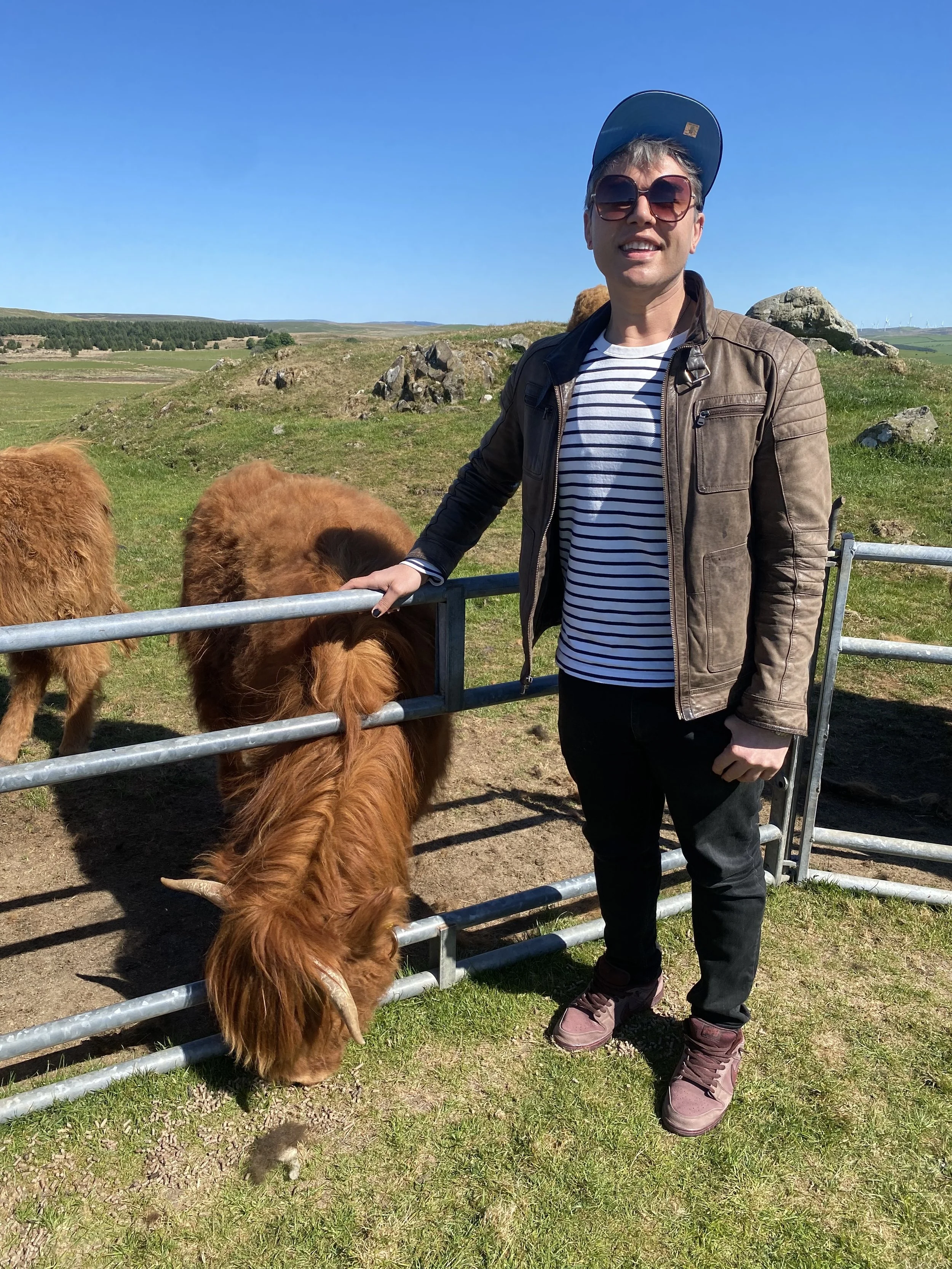A person wearing sunglasses, a baseball cap, a leather jacket, and striped shirt beside a cow on a farm besides rolling hills and a clear blue sky.