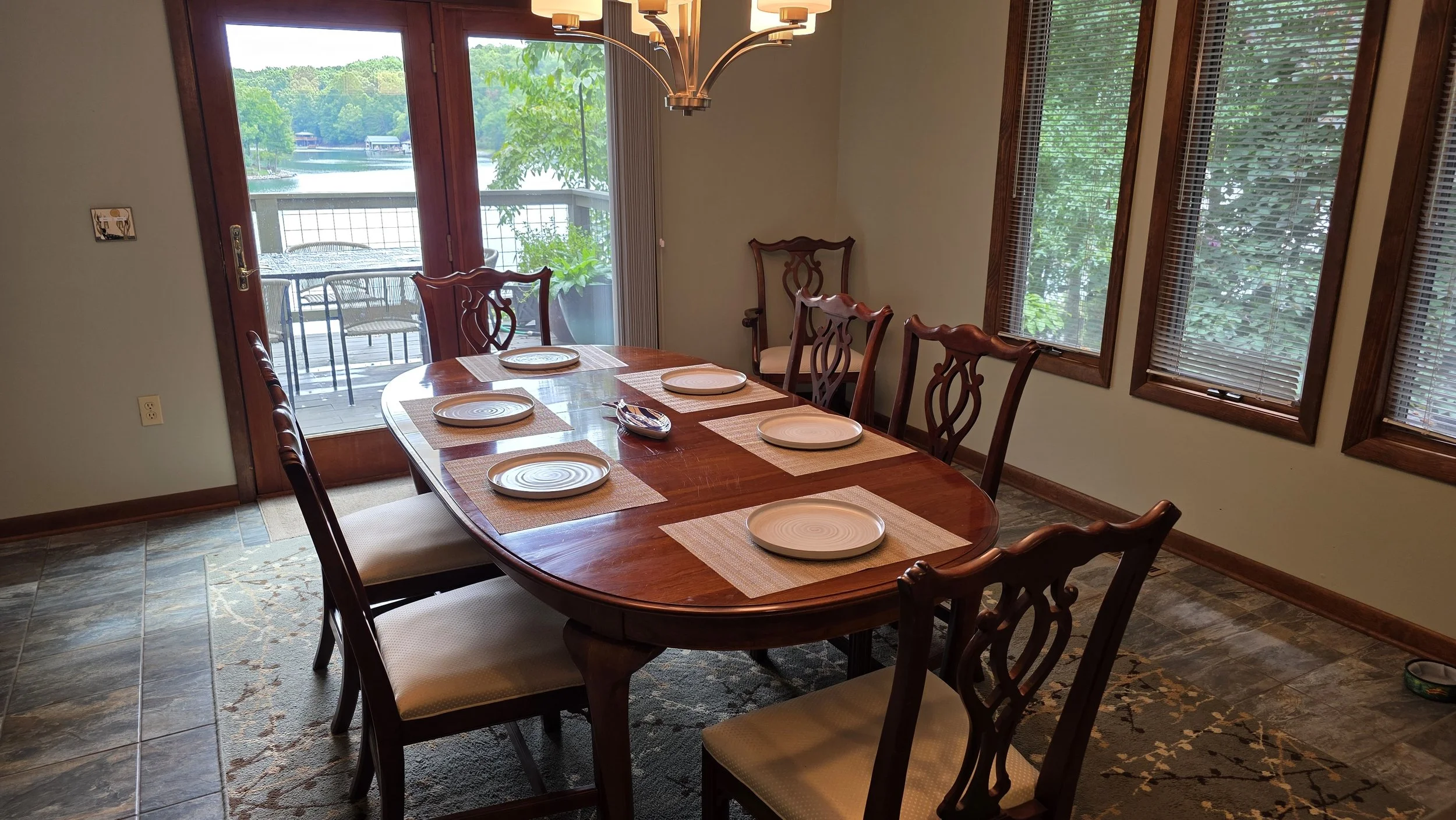 A dining room with a wooden table set for six, featuring beige placemats and white plates, with a view of a lake and trees through sliding glass doors and windows.