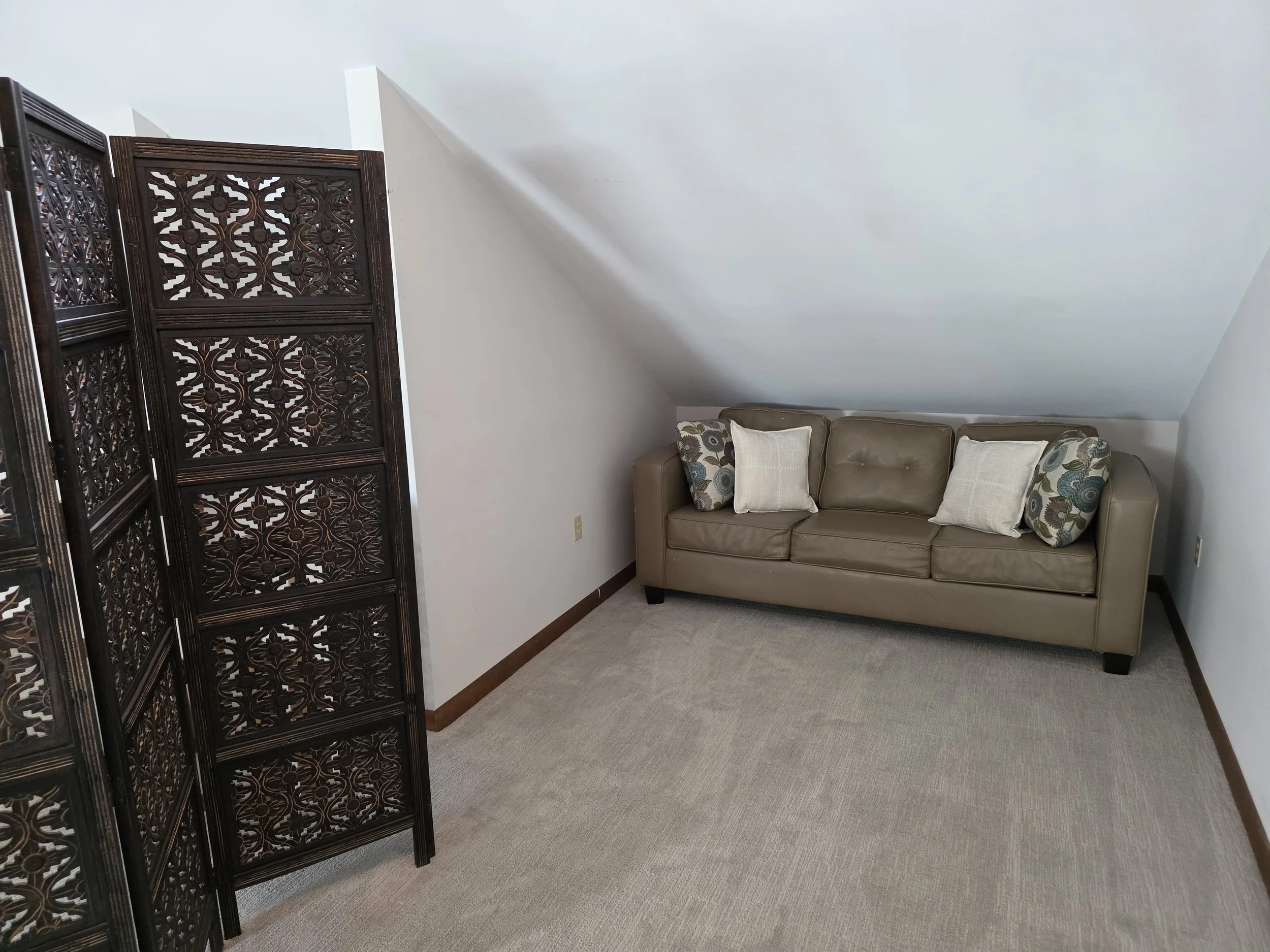 Living room with a beige sofa and decorative pillows, light-colored carpet, and a folding wooden room divider with intricate lattice design, under a sloped ceiling.