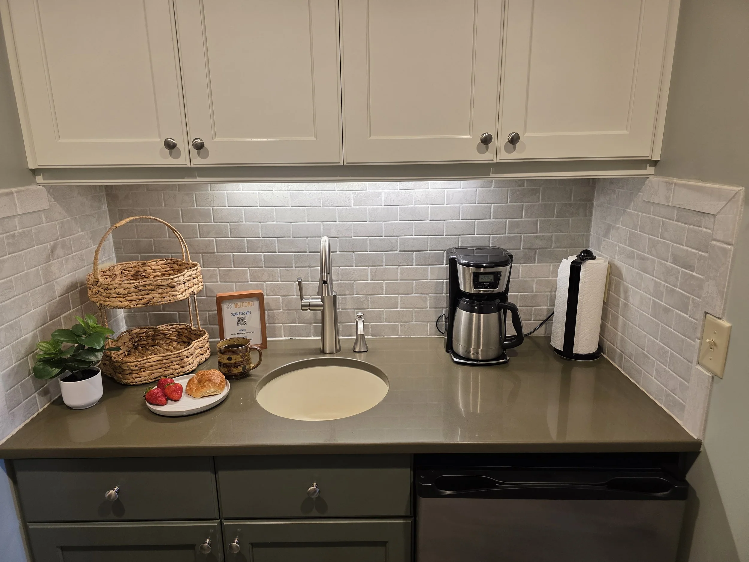 Kitchen countertop with coffee maker, paper towel holder, plant, strawberries, croissant, coffee mug, wicker baskets, and decorative sign, with beige cabinets and gray backsplash.