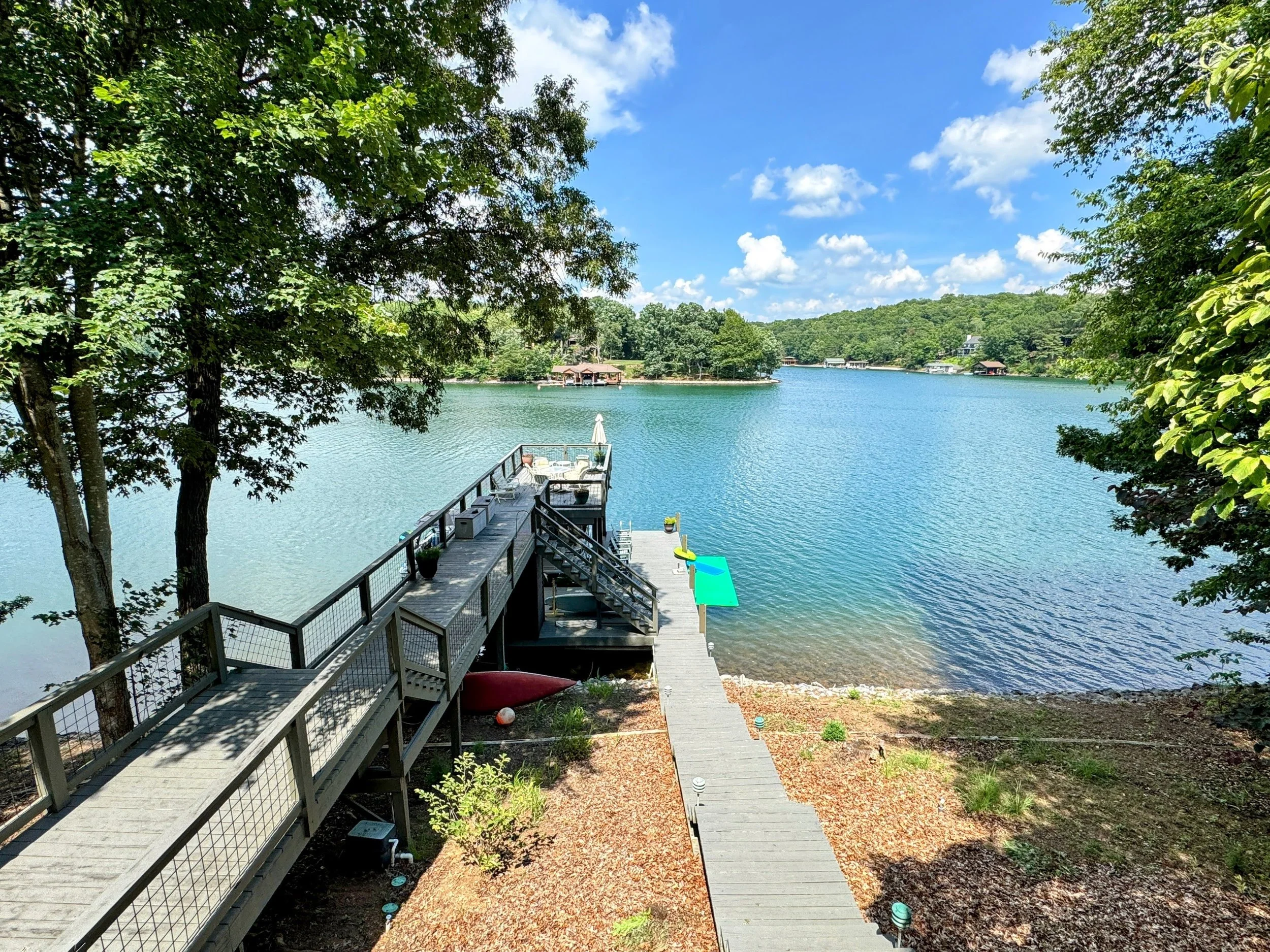 View of a lake with a wooden dock and stairs leading into the water, surrounded by trees under a partly cloudy sky.