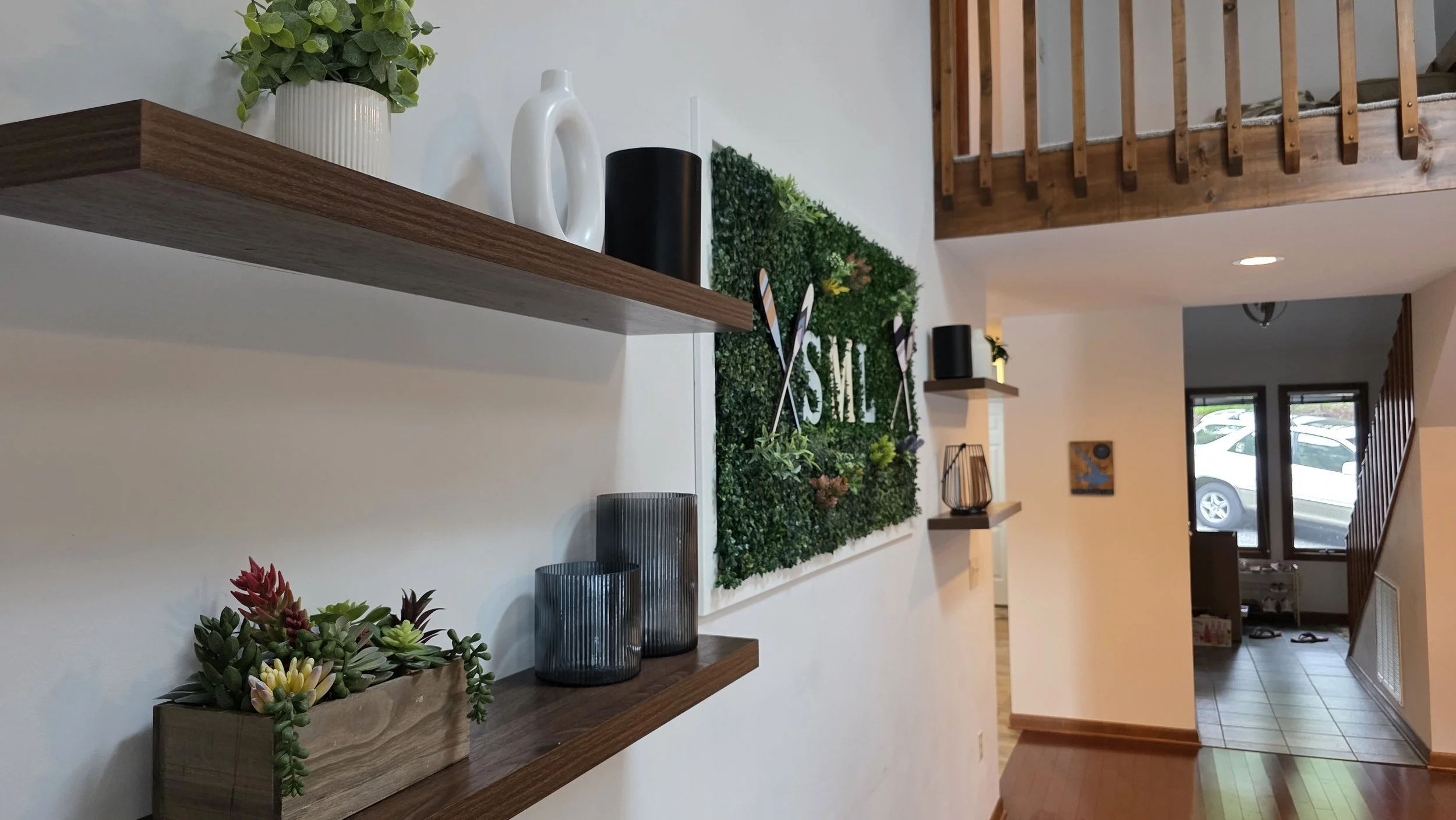 Living room entryway with two wooden shelves decorated with plants and vases, a green wall art piece with the word 'SIX', and a staircase leading to an upper level. The area has a front window showing parked cars outside.