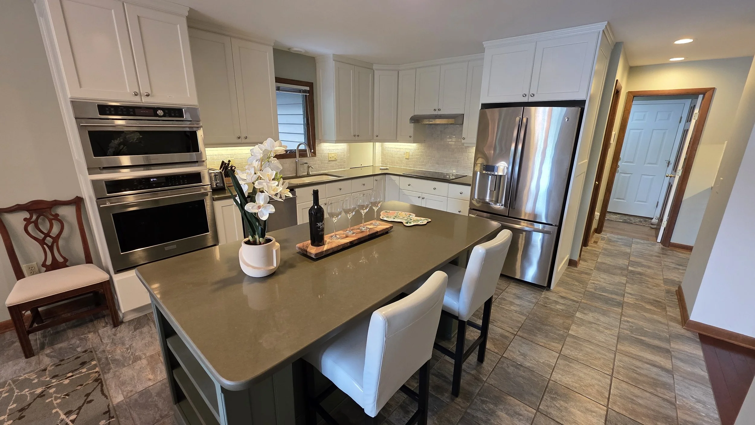 Modern kitchen with white cabinets, stainless steel appliances, and a central island with white chairs. Decor includes a potted orchid, a bottle of wine, and four glasses, with sunlight coming through a window.