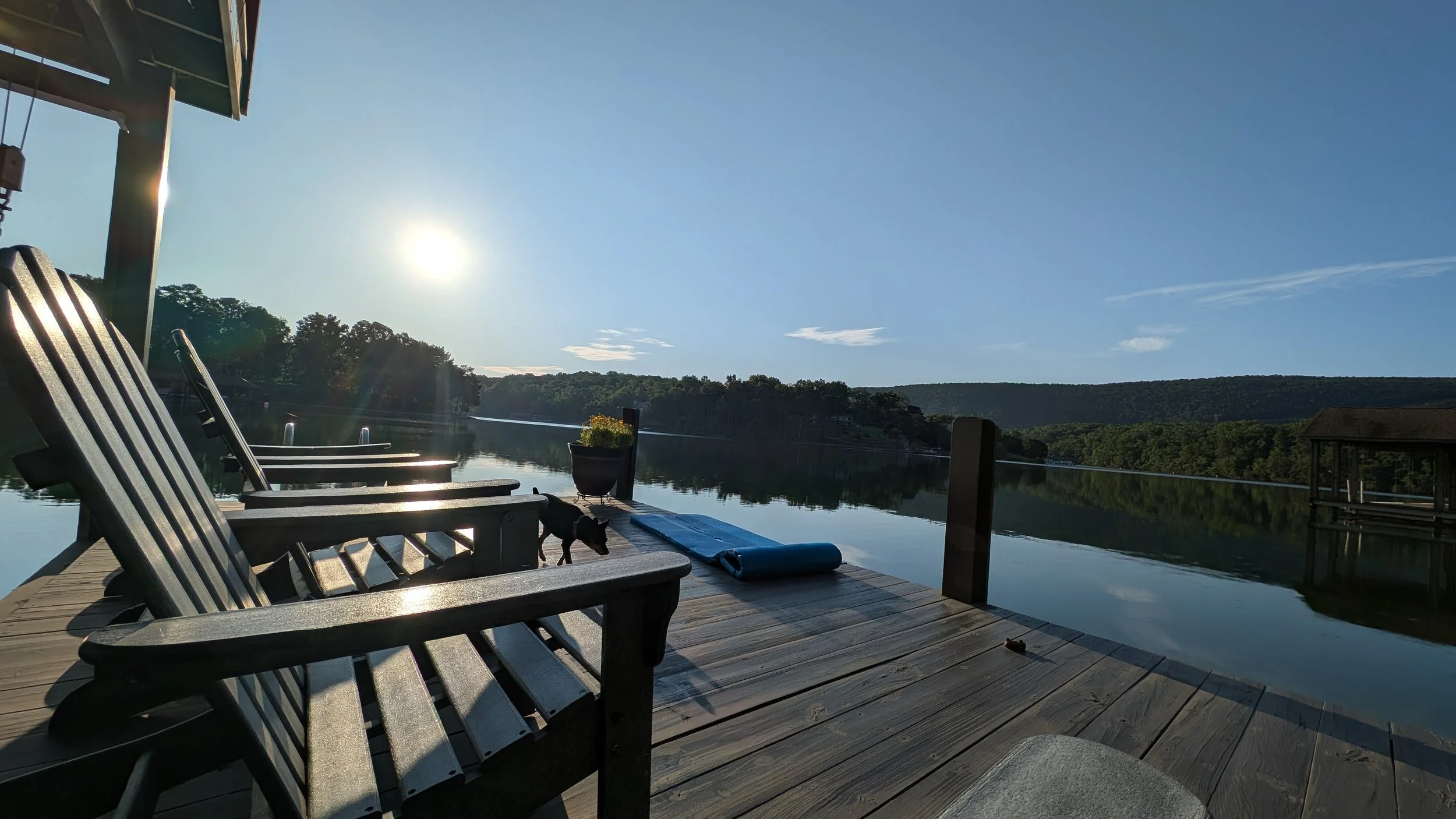 A lakeside dock with two wooden chairs, a rolled-up yoga mat, and a small dog near a potted plant. The water is calm, reflecting the trees and sky, with the sun low in the sky.