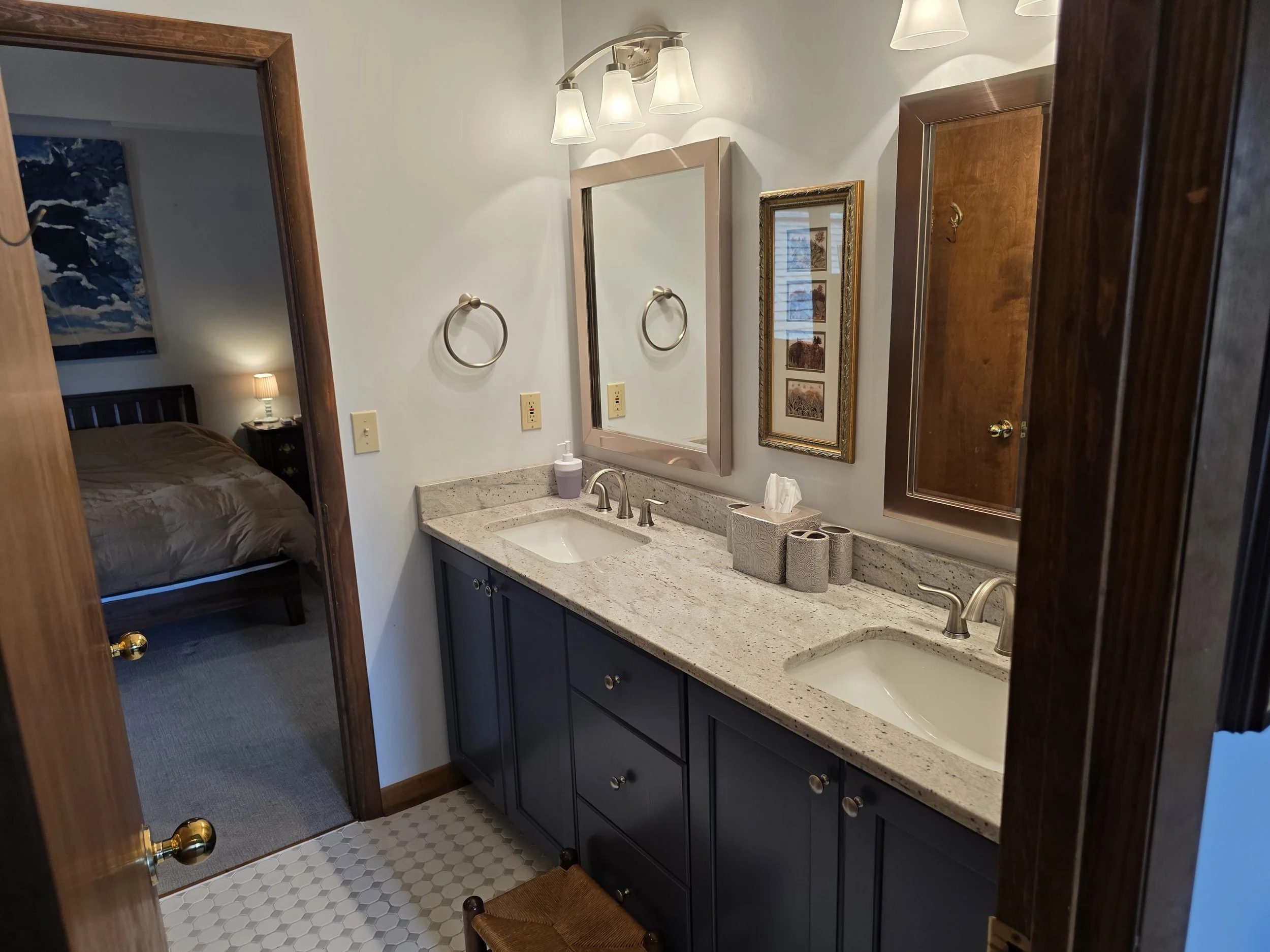 Bathroom with double sink vanity, framed mirror, wall-mounted lights, bedroom visible through doorway, and decorative framed picture on wall.