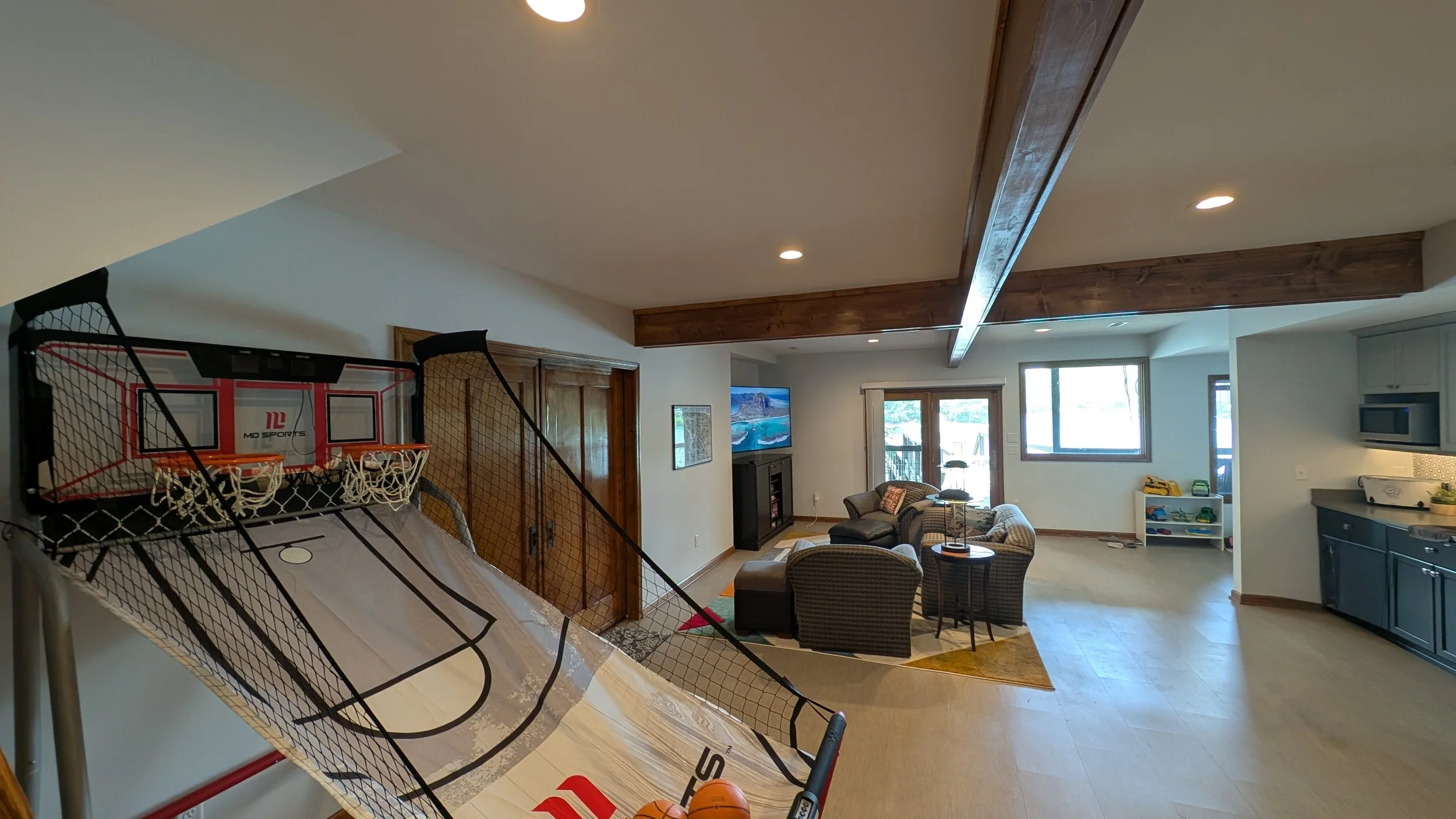 Living room with basketball arcade game, seating area with chairs and sofa, television, windows, and kitchen area.