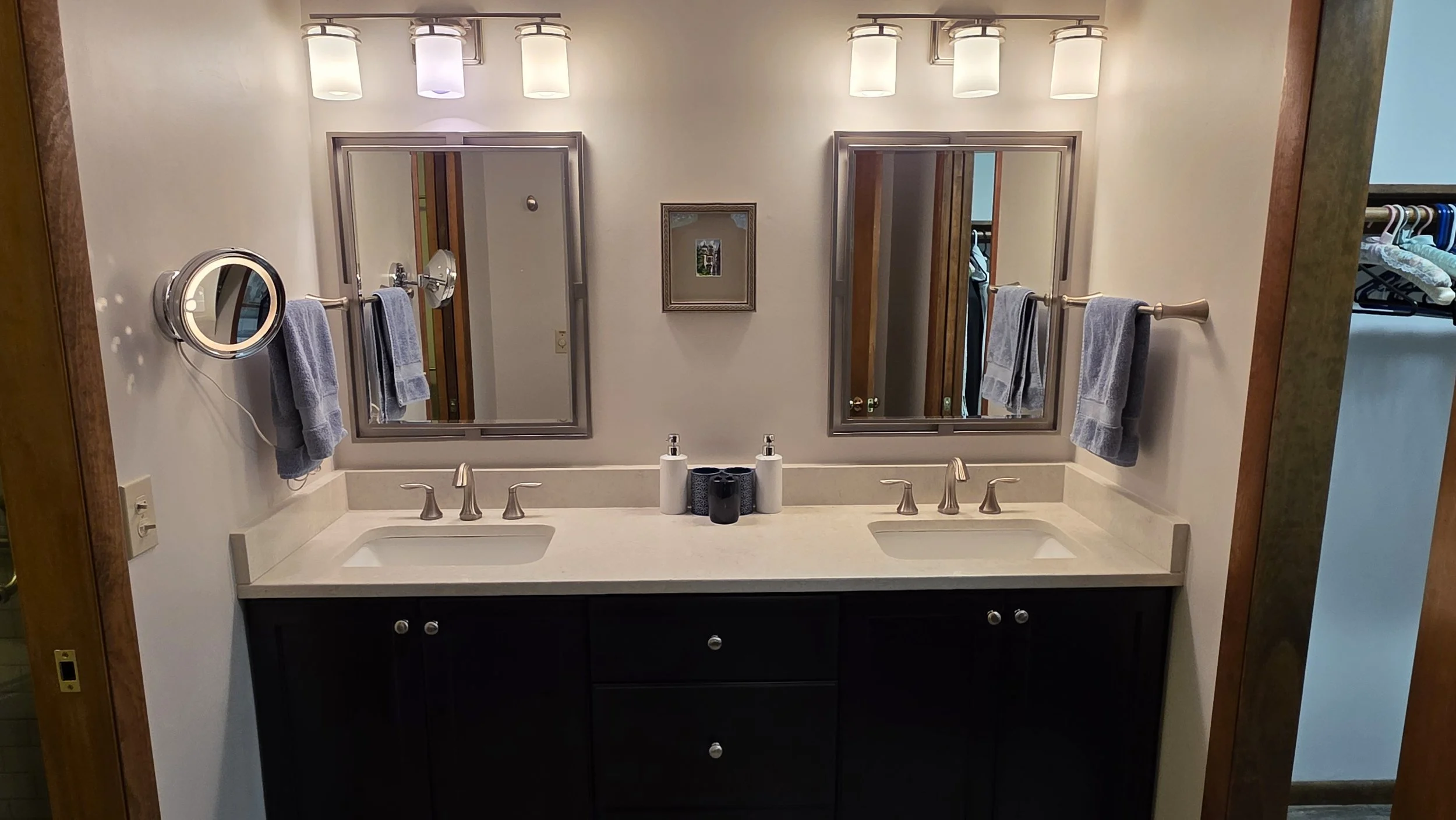 Double bathroom vanity with two mirrors, two sinks, and four light fixtures above. Each side has a towel bar with a gray towel. Small framed picture between the mirrors. Items on the countertop include bottles of soap or lotion and a black container.