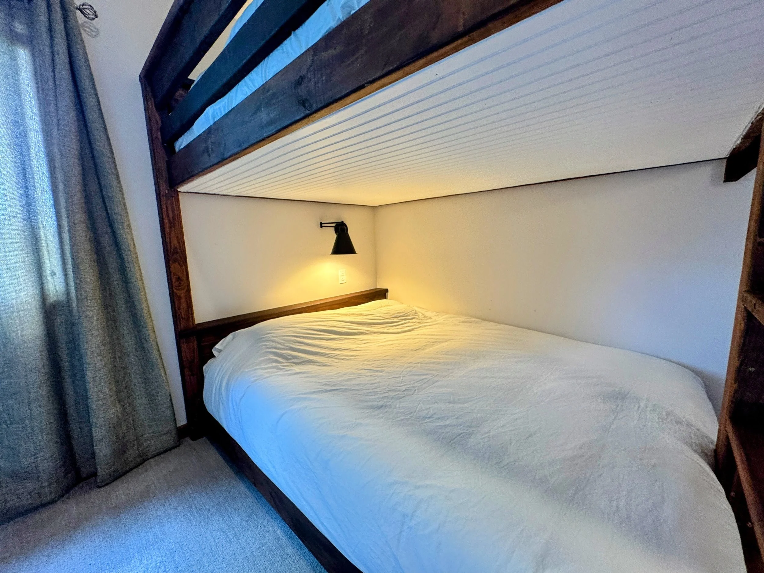 Bedroom with a single bed and a lofted bed above it, featuring wooden framing, a bedside wall lamp, beige bedding, gray curtains, and neutral-colored carpet.