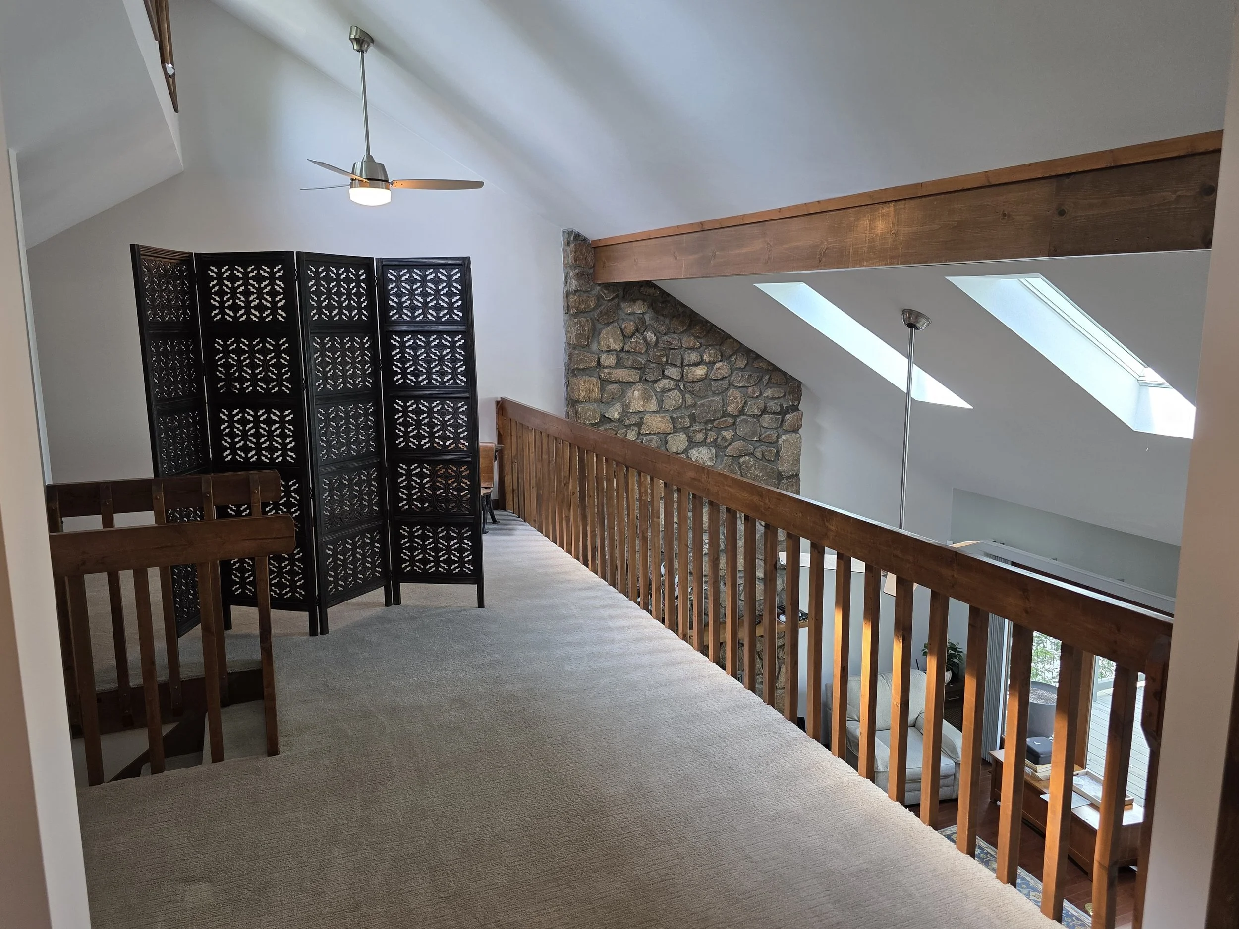 Upper level of a house with a wooden railing, black decorative room divider, ceiling fan, skylights, and exposed stone wall.