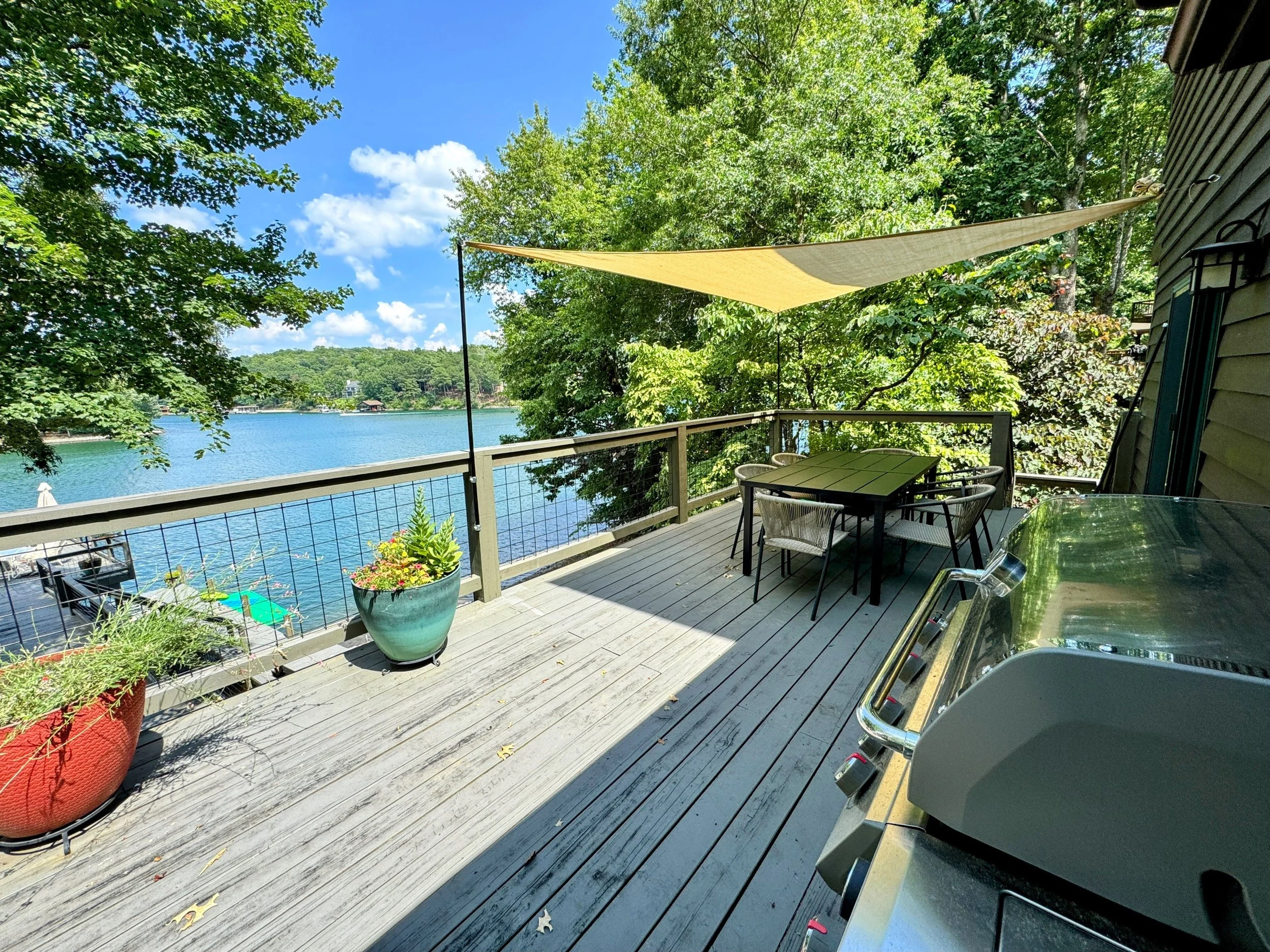 A spacious outdoor deck overlooking a lake, with a table and chairs, potted plants, a sunshade, and trees surrounding the area.