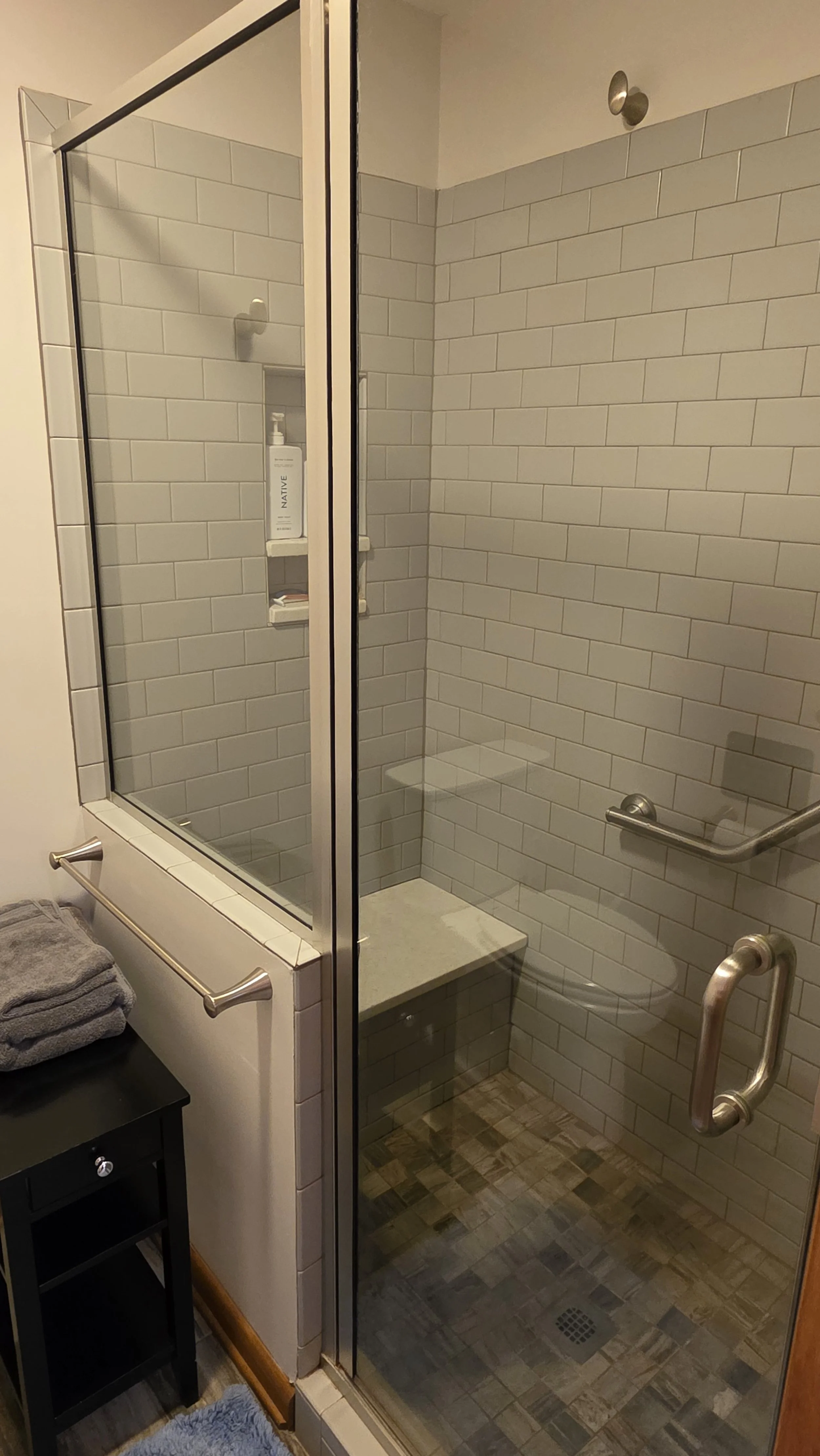 A walk-in shower with beige subway tiles, a bench, a grab bar, and a glass door, with a small black side table holding gray towels nearby.