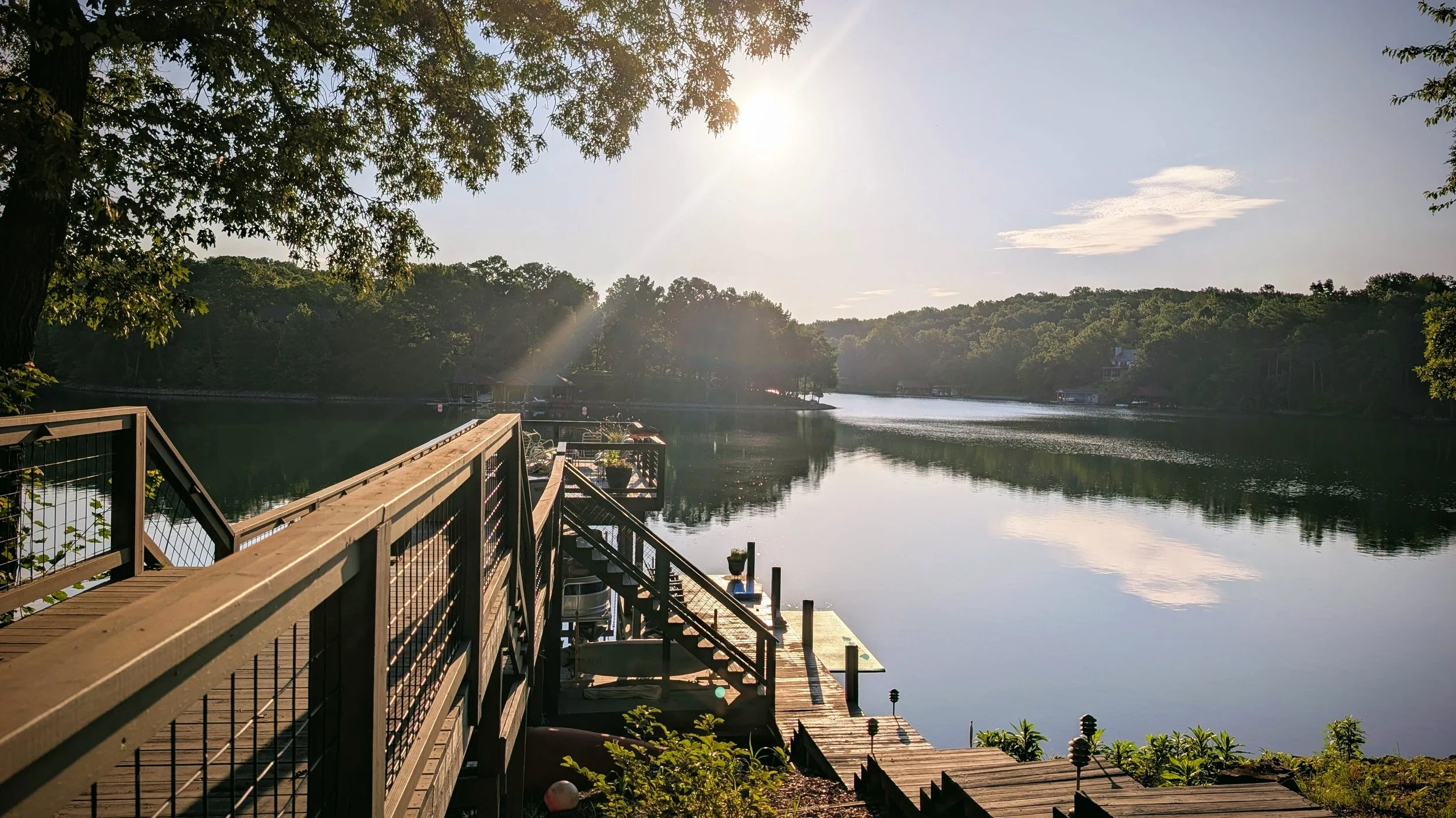 A wooden dock with railings extends over a calm river, reflecting the sky and nearby trees. The sun is shining brightly, creating a warm glow over the greenery and water.