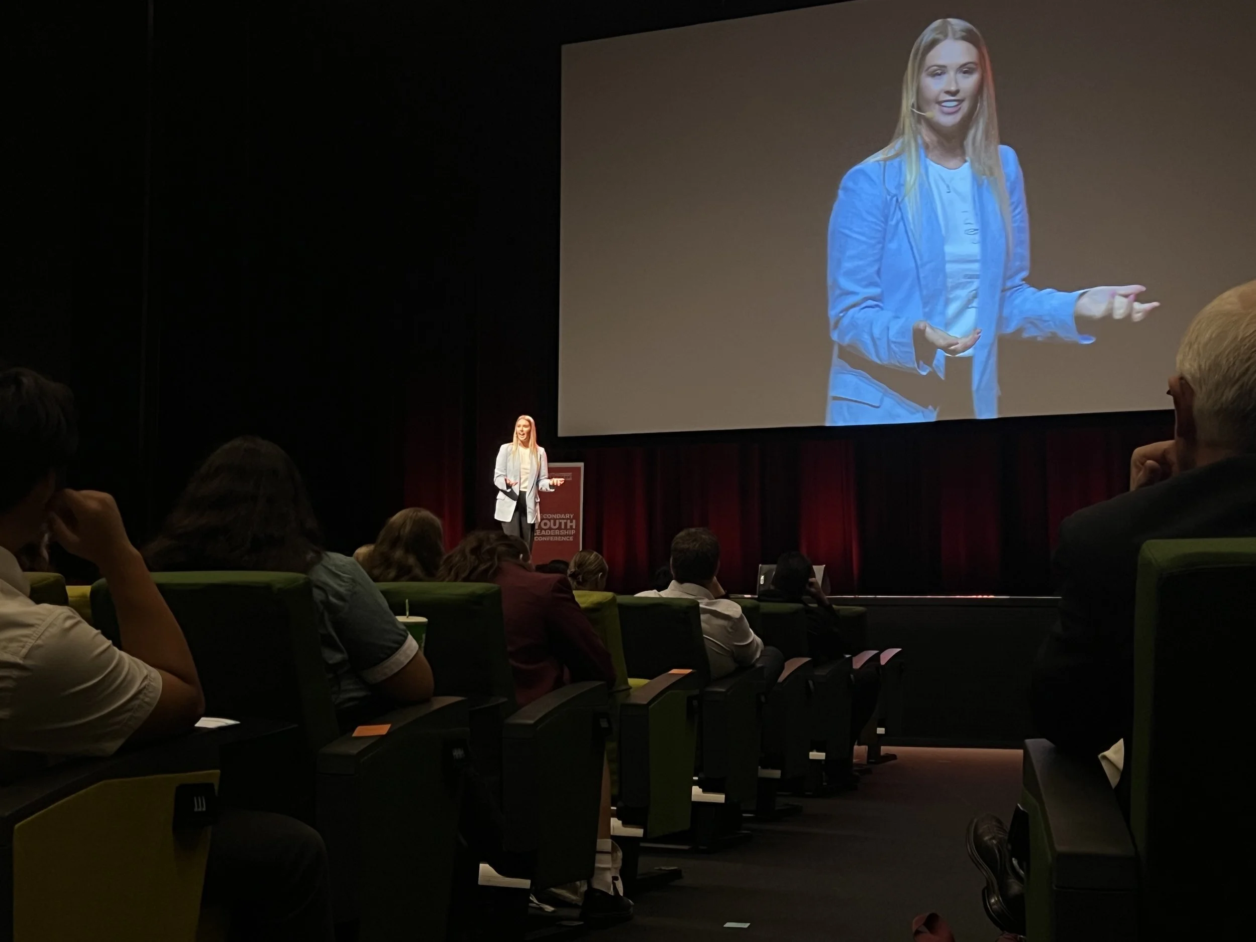 A woman giving a presentation on stage at a conference, seen on a large screen behind her, with an audience seated in rows watching.