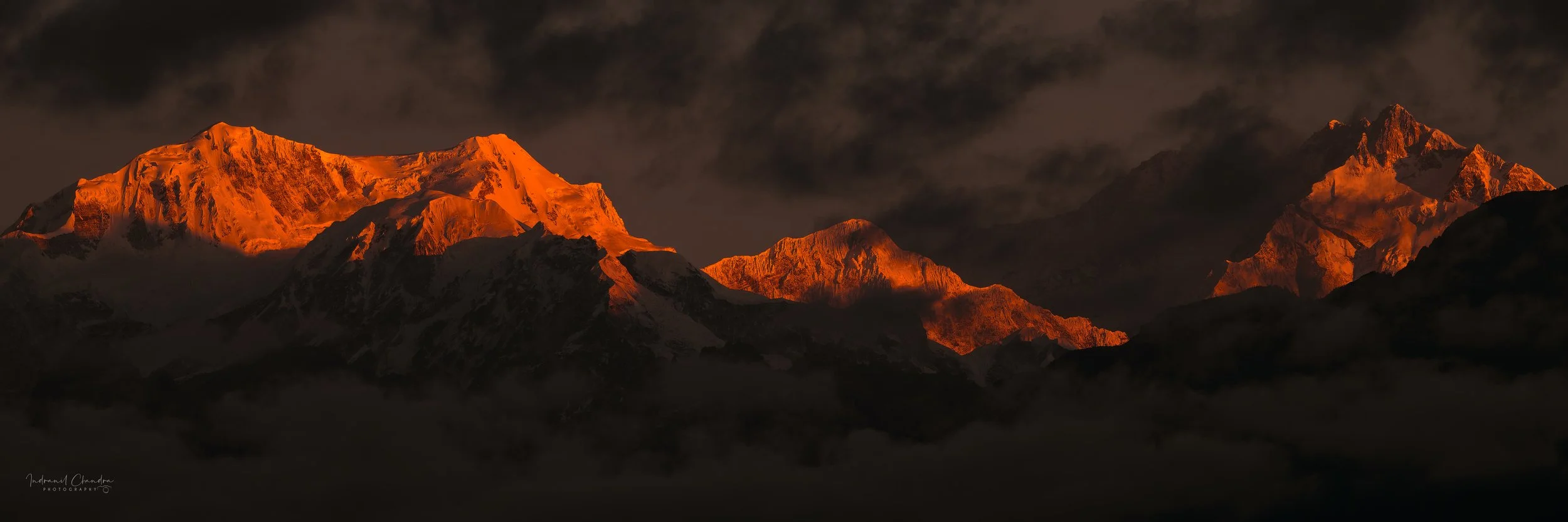 Sunset illuminates snow-capped mountain peaks with an orange glow, with dark clouds in the sky.