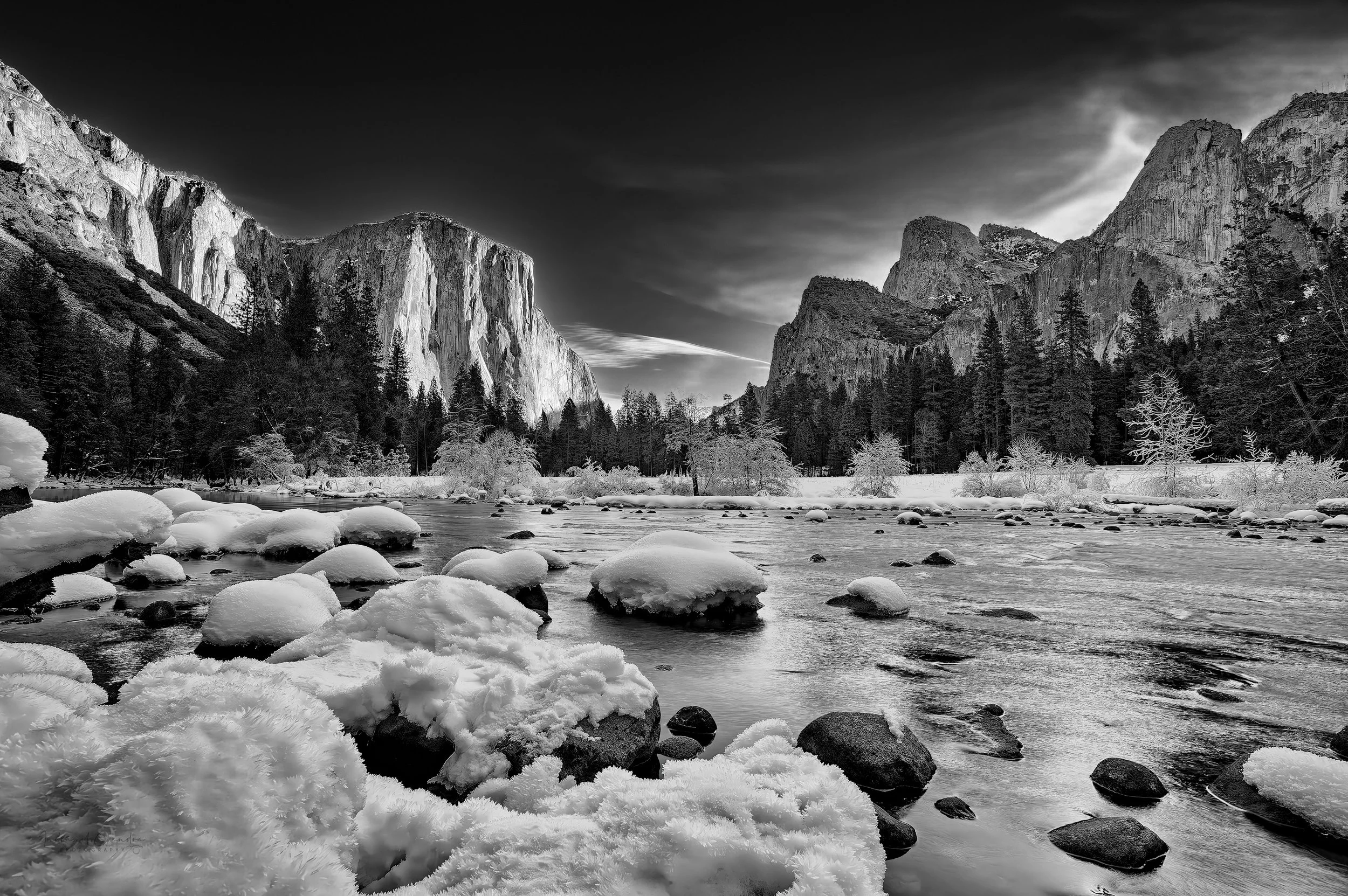 A black and white photo of snow-covered rocks along a river with trees and mountain cliffs in the background under a cloudy sky.