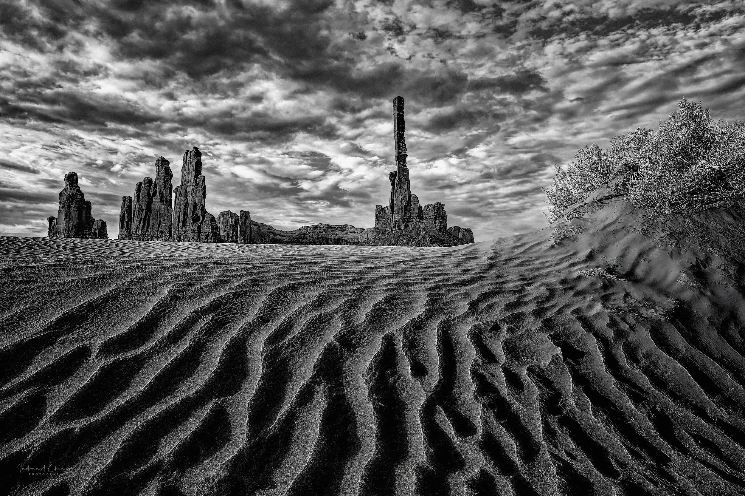 Black and white photograph of a desert landscape with textured sand and rocky formations in the background, cloudy sky overhead.