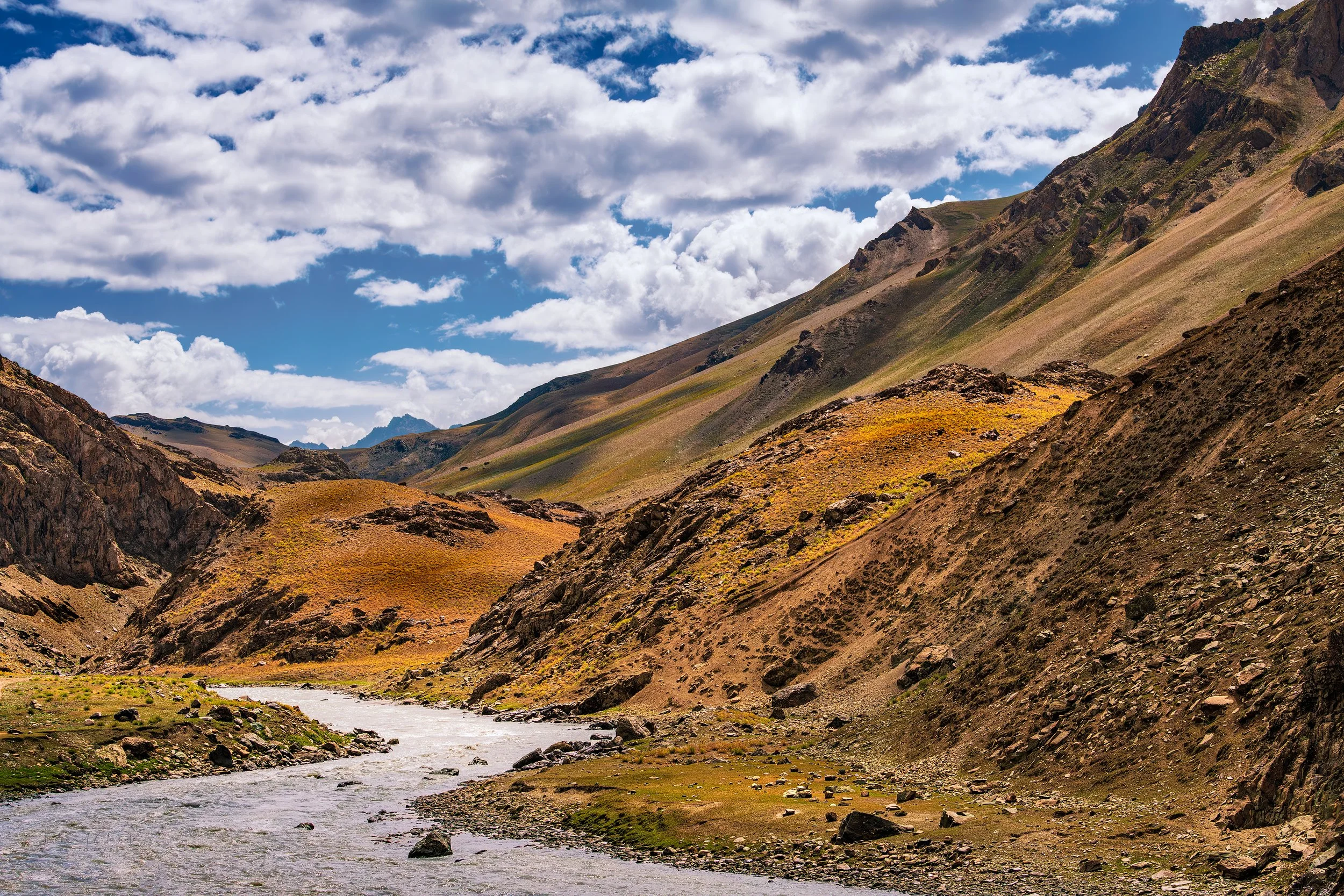 Mountain stream meandering through the colorful slopes of a Kashmir valley, where rugged terrain meets open skies and scattered clouds.