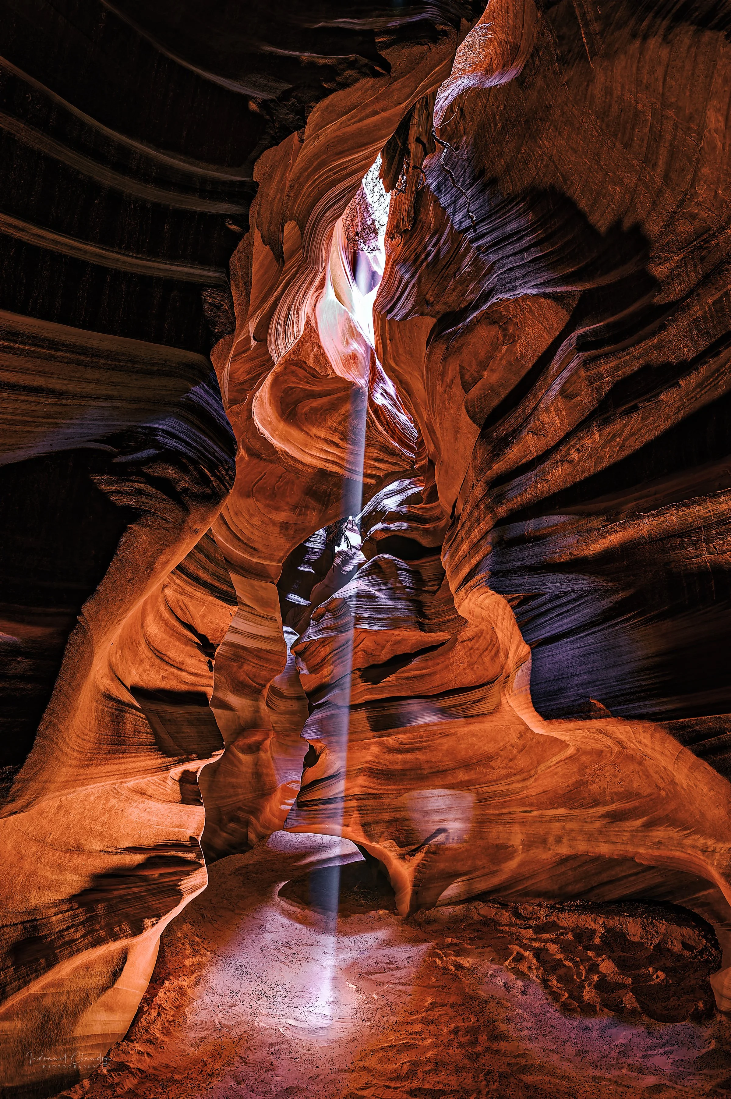 A solitary beam of light illuminates the flowing, wave-like sandstone walls of Antelope Canyon, creating a dramatic contrast of light and shadow and highlighting the vibrant orange, red, and purple hues of the rock formations. This ethereal "God beam