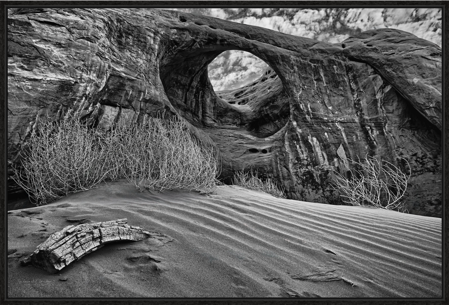 Black and white photograph of a desert landscape with a large rock formation featuring a natural circular arch, sandy dunes with ripples, dry bushes, and a piece of wood on the sand, under a cloudy sky.