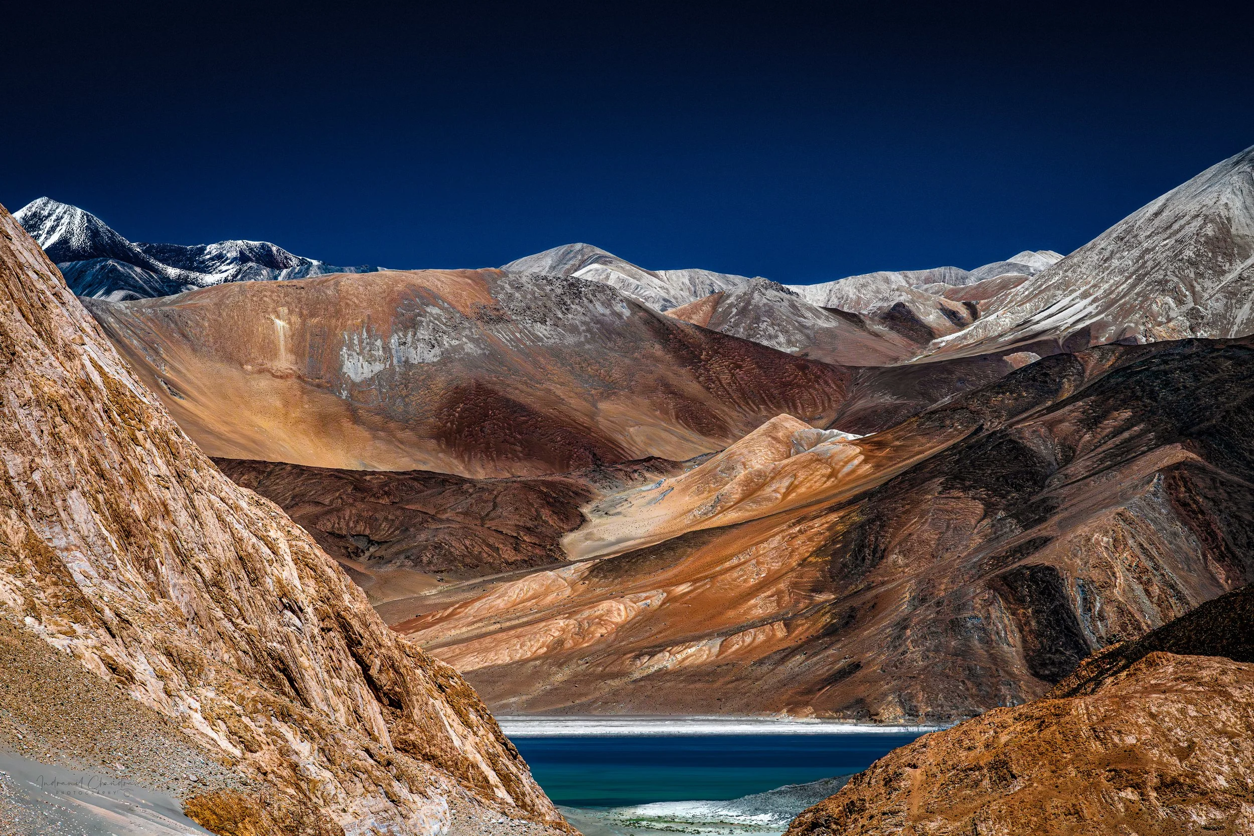 Colorful mountain landscape with snow-capped peaks, rocky slopes in various shades, and a blue lake in the foreground under a clear dark sky.