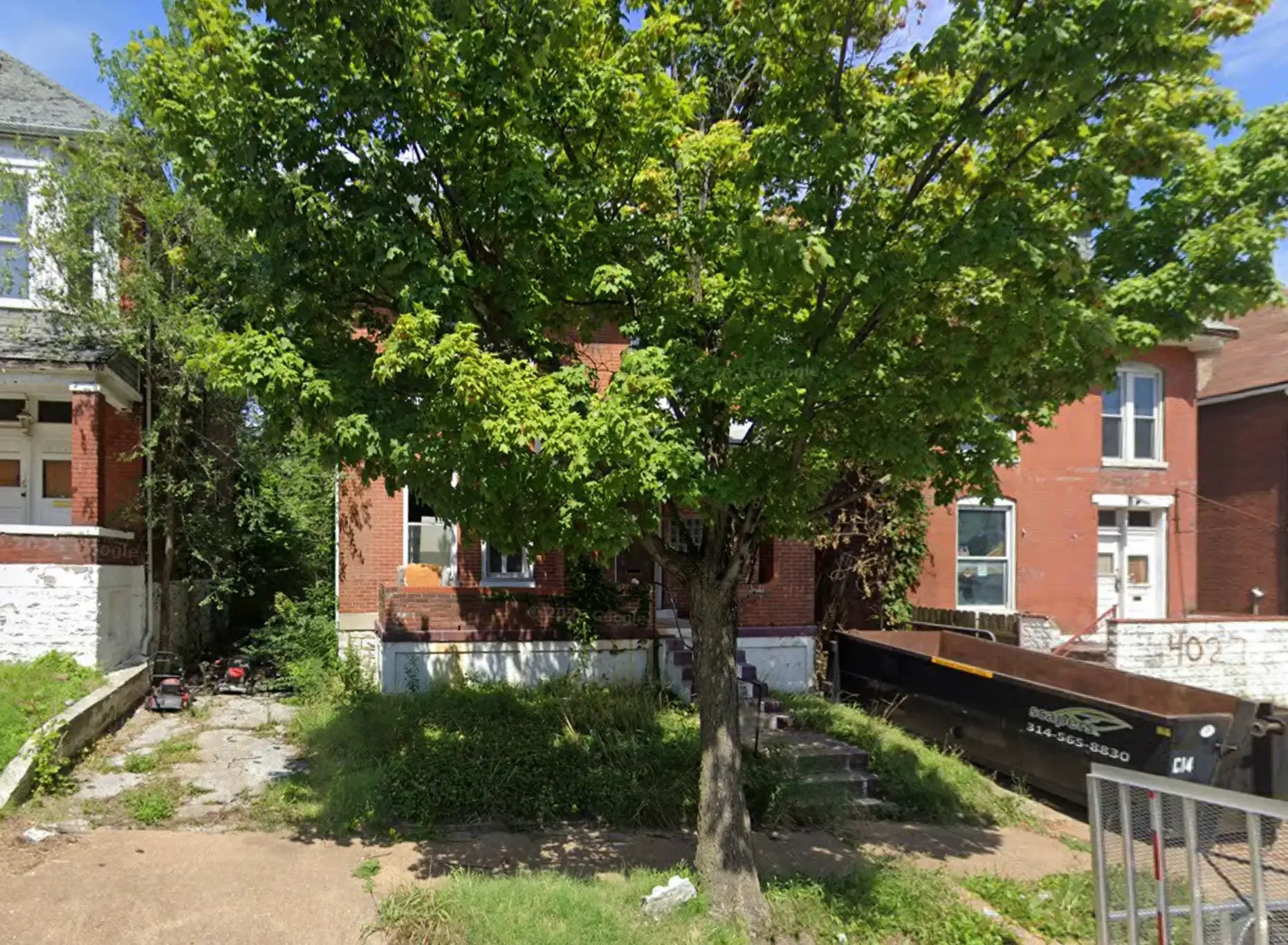 A large green tree in front of a red brick house with white trim. The house has steps leading to a door and windows. A black dumpster is on the right side, and there is some grass and a sidewalk in the foreground.