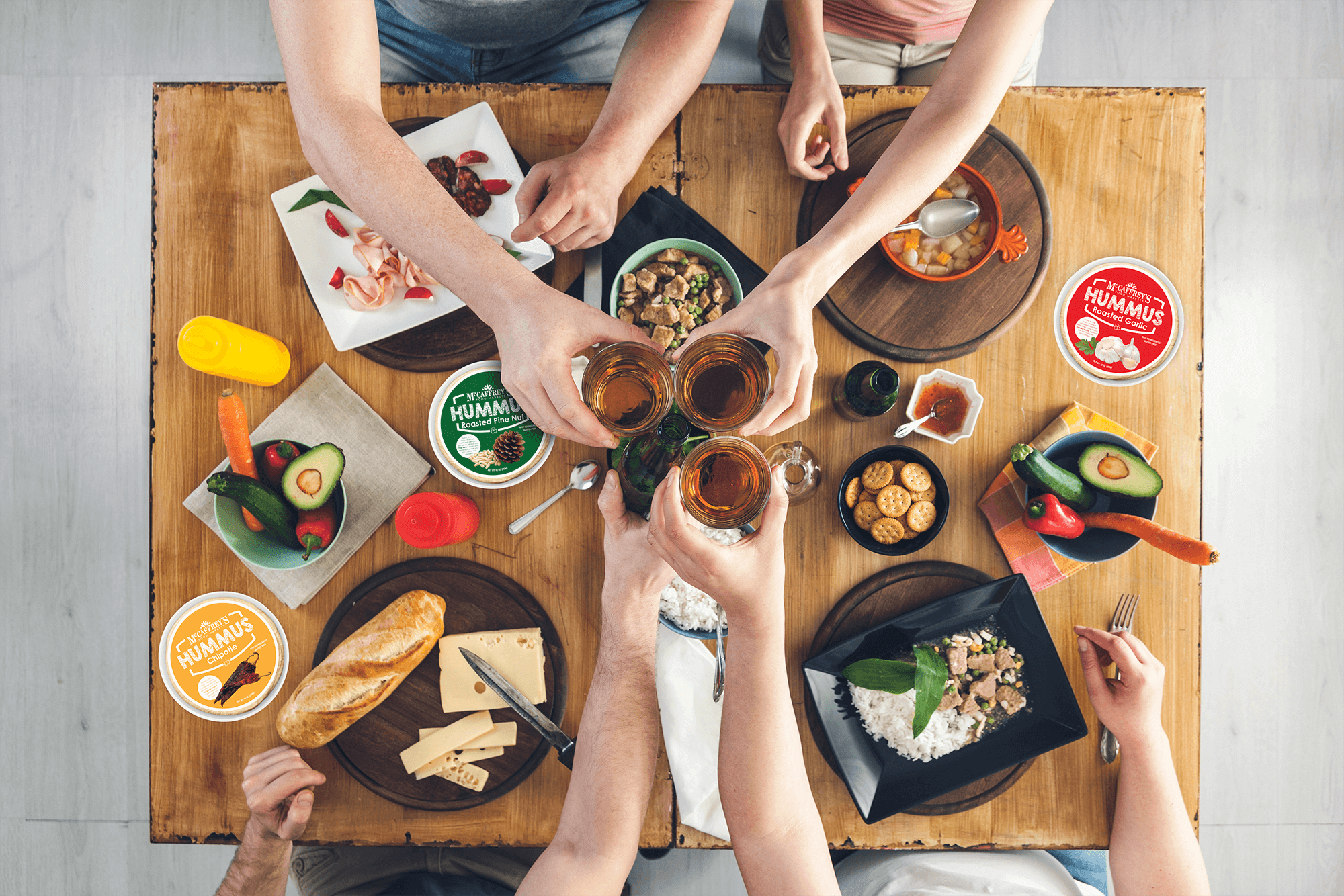 Overhead view of people cheering glasses with McCaffrey's hummus, bread and fresh vegetables on wooden table
