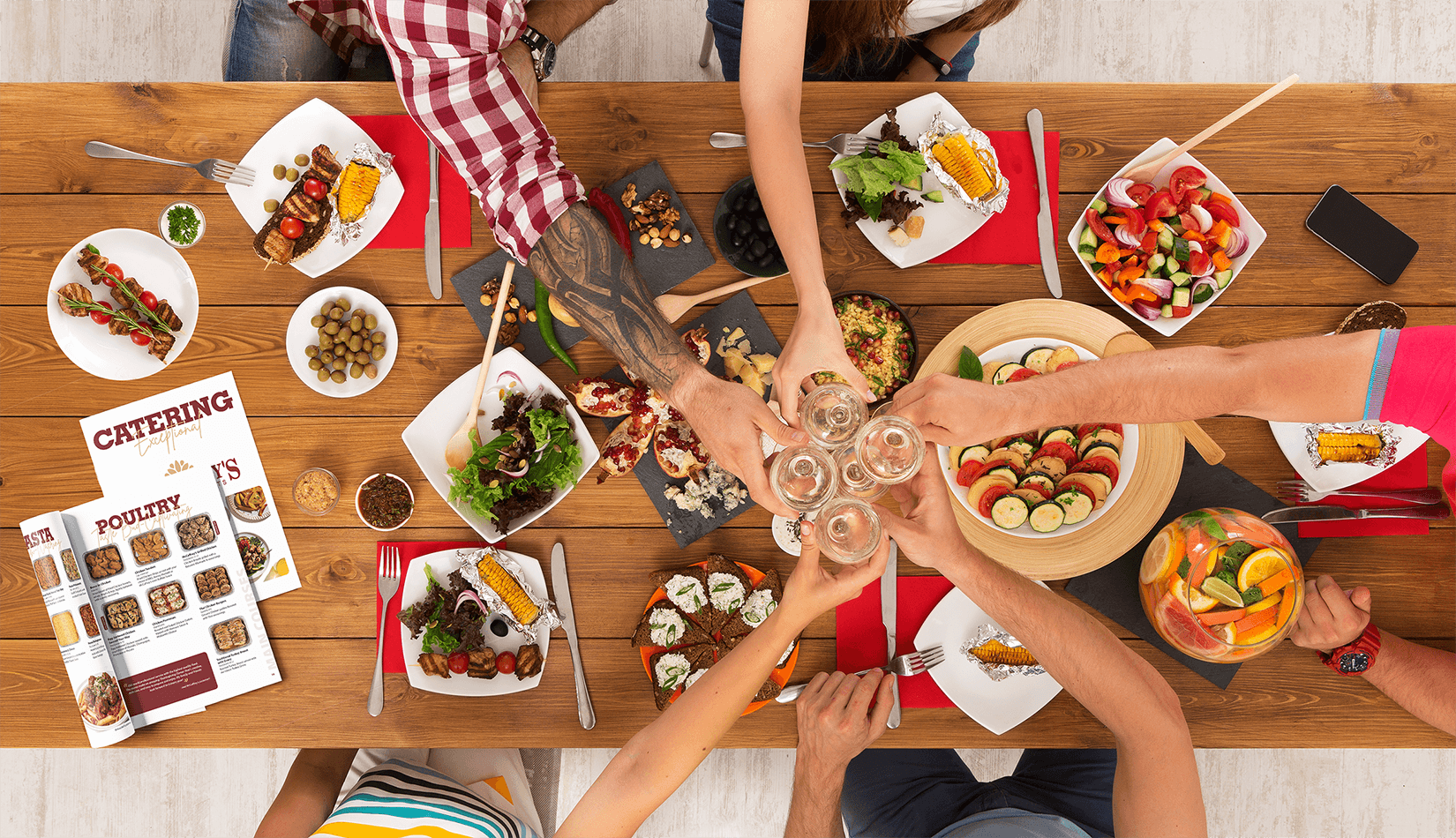 Overhead shot of people cheering glasses with food and two copies of the McCaffrey's catering book