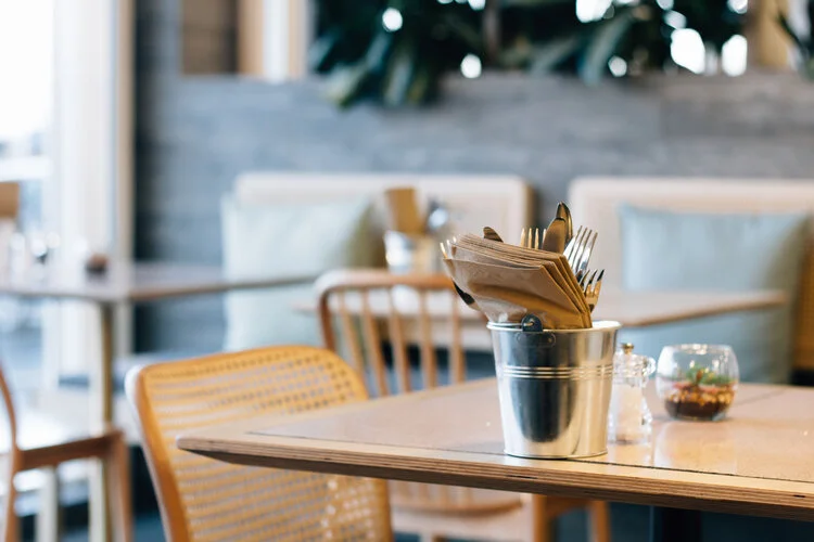 A table in a restaurant with a metal container holding paper napkins and utensils, and a small glass bowl with a succulent plant, in a cozy dining area with wooden chairs and booth seating.