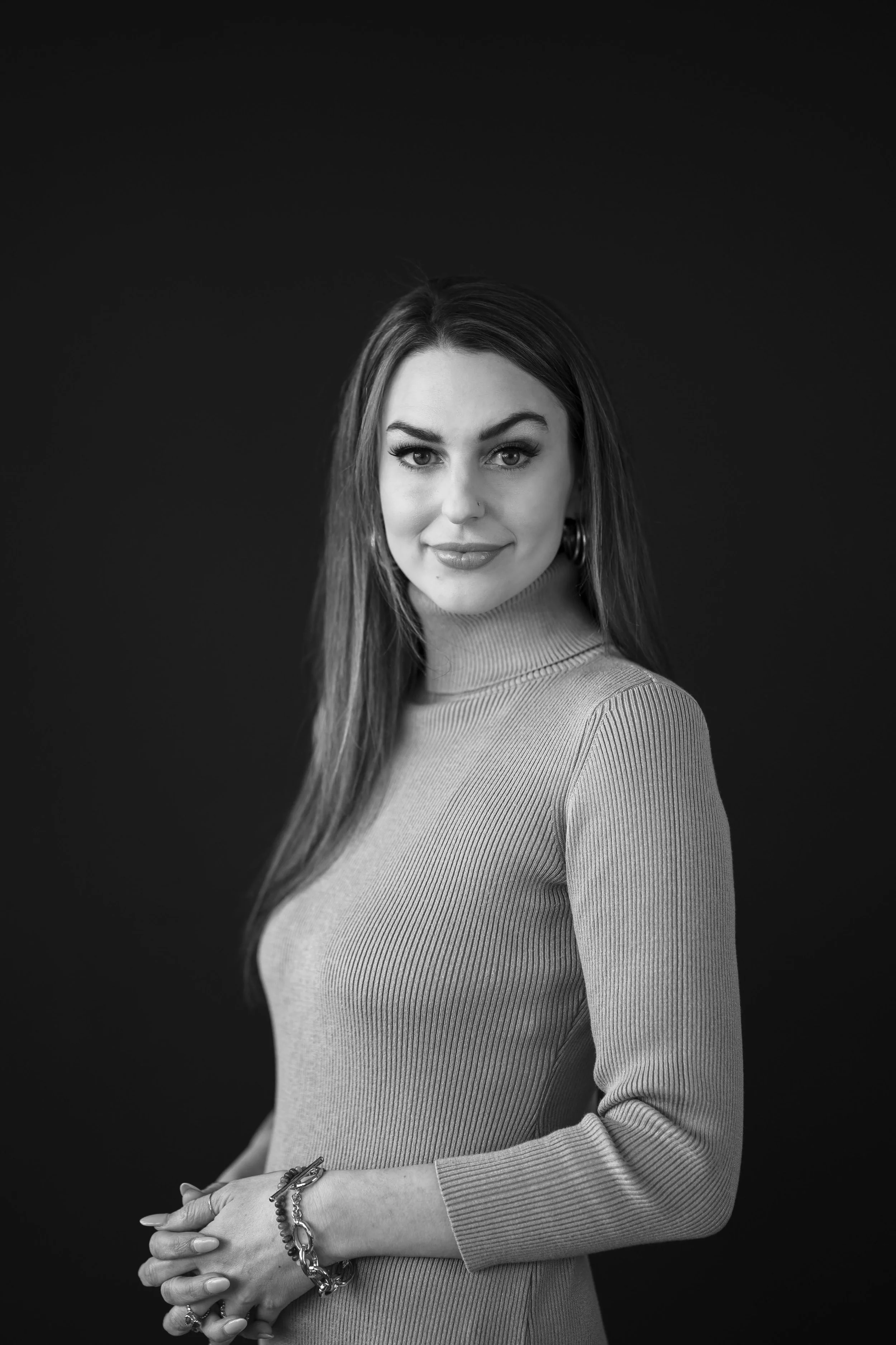 Black and white portrait of a woman with long hair, wearing a ribbed turtleneck sweater, with a slight smile and jewelry on her wrist and fingers, against a dark background.