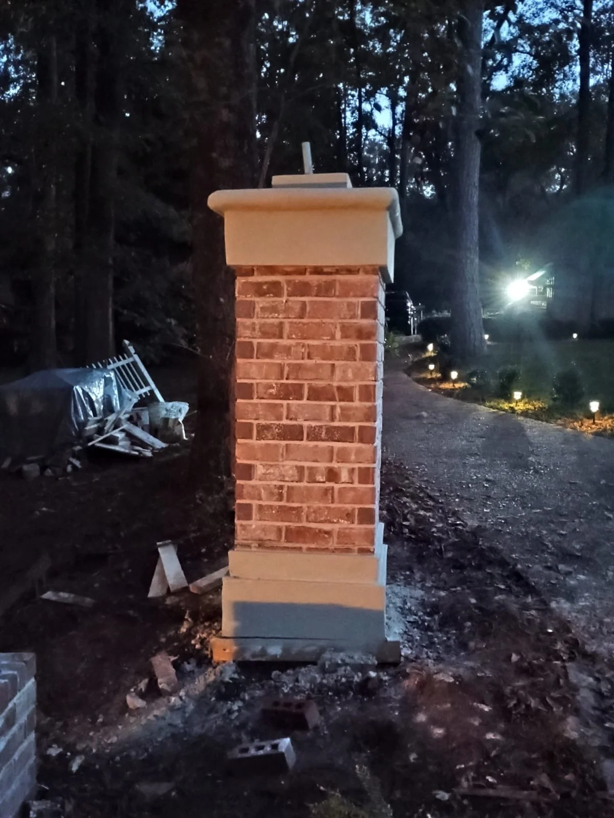 Una columna de ladrillos con remate de cemento en un área exterior durante la noche, con árboles y sendero iluminado en el fondo.