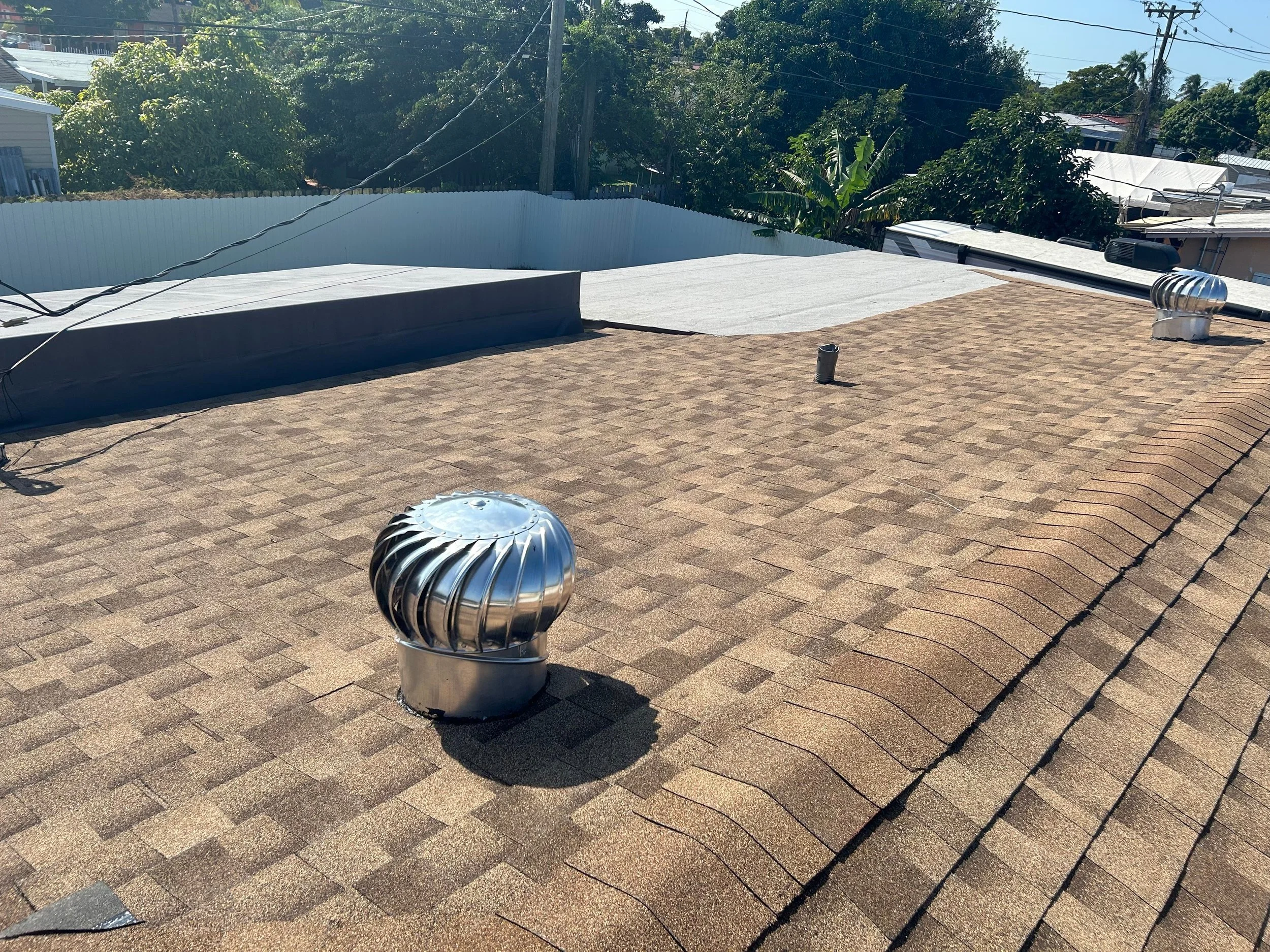 Techo de tejas de asphalt con varios respiraderos metálicos en una vivienda, con árboles y vivienda en el fondo.