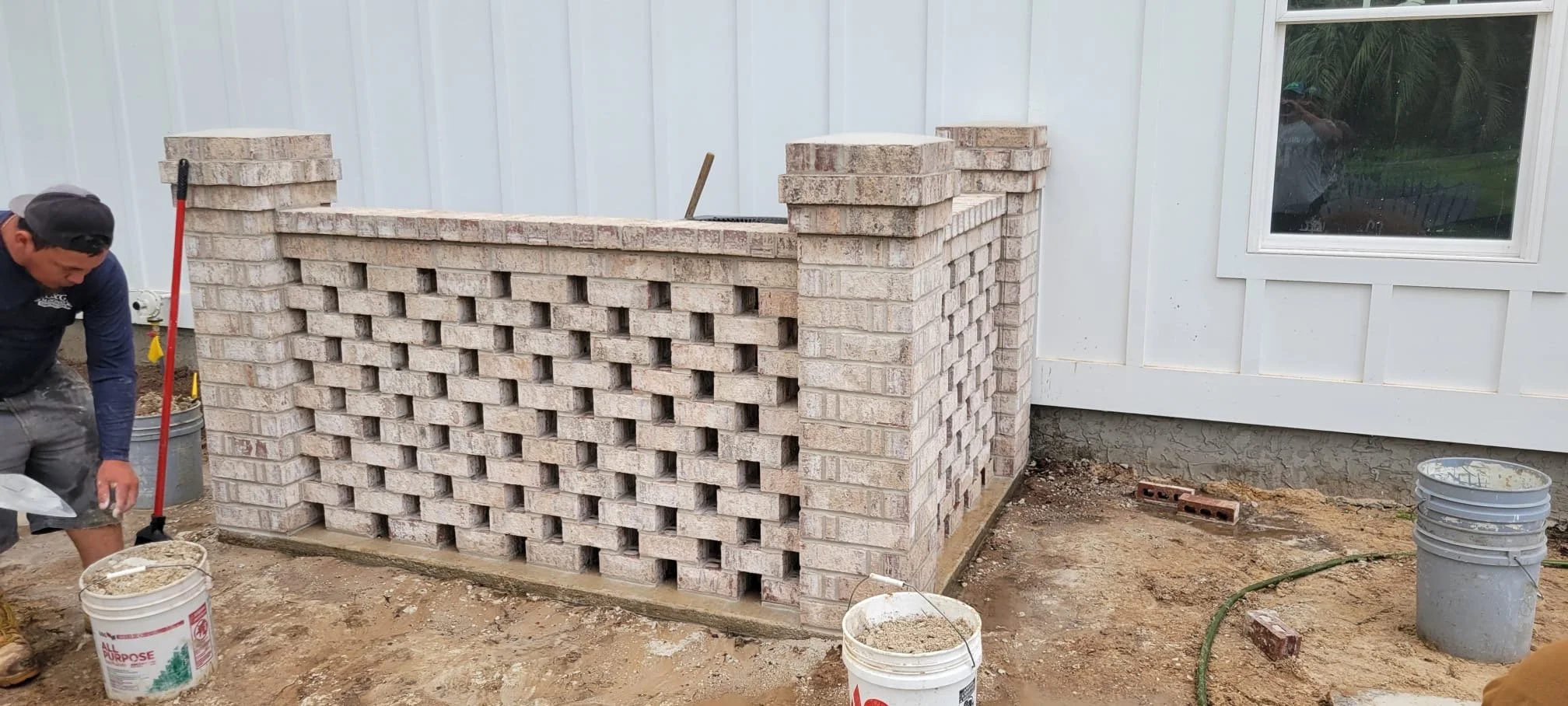 Construcción de un muro de bloques de ladrillo frente a una pared de una casa blanca, con un hombre trabajando y cubetas con mezcla de cemento en el suelo.