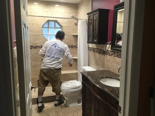 Hombre de espaldas limpiando el inodoro en un baño. Tiene puesto un uniforme de limpieza y está usando guantes.