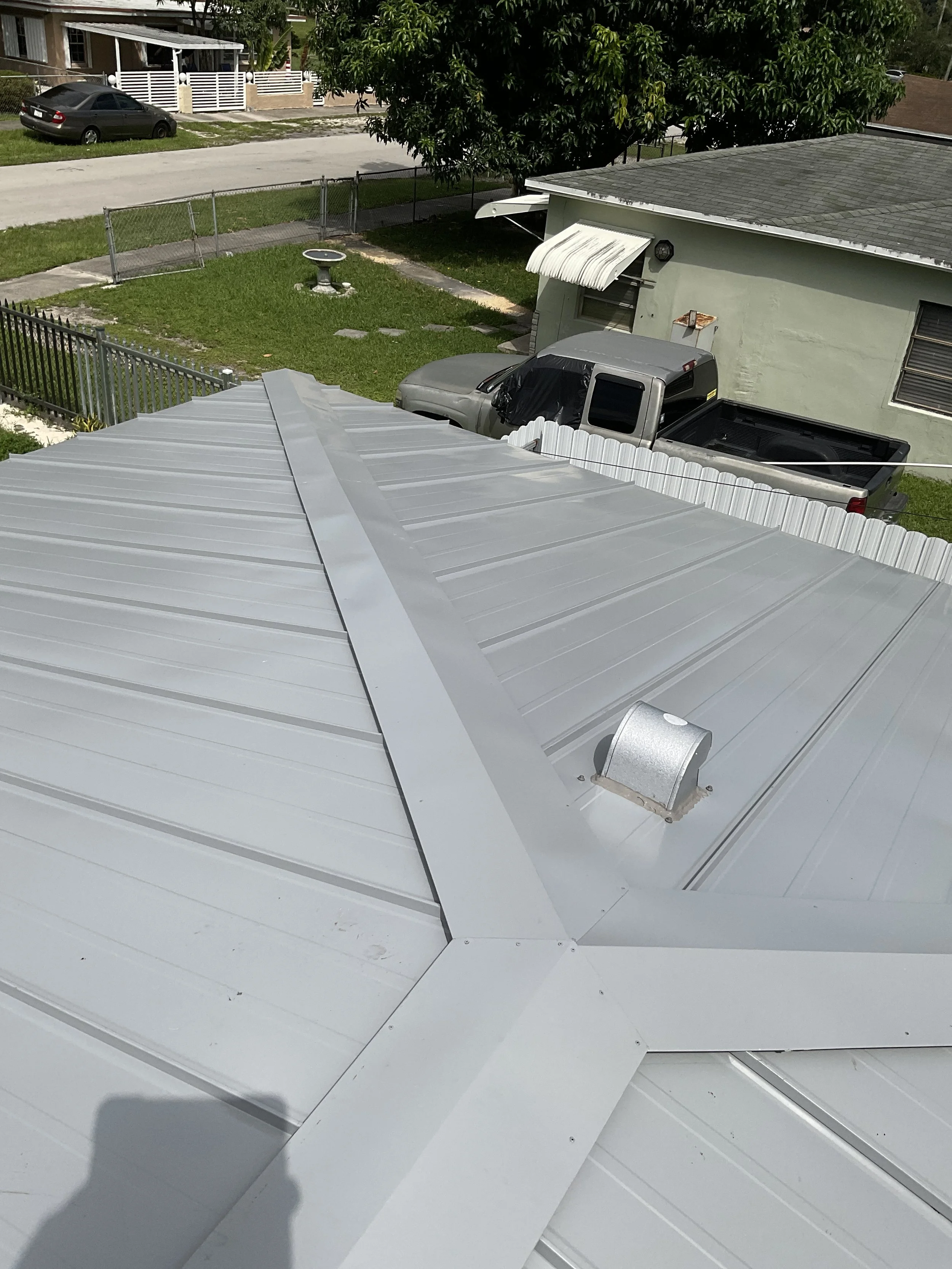Vista desde arriba de un techo de metal gris con una pequeña ventilación, con casas, árboles, un automóvil, y un camión en el fondo, en una zona residencial.