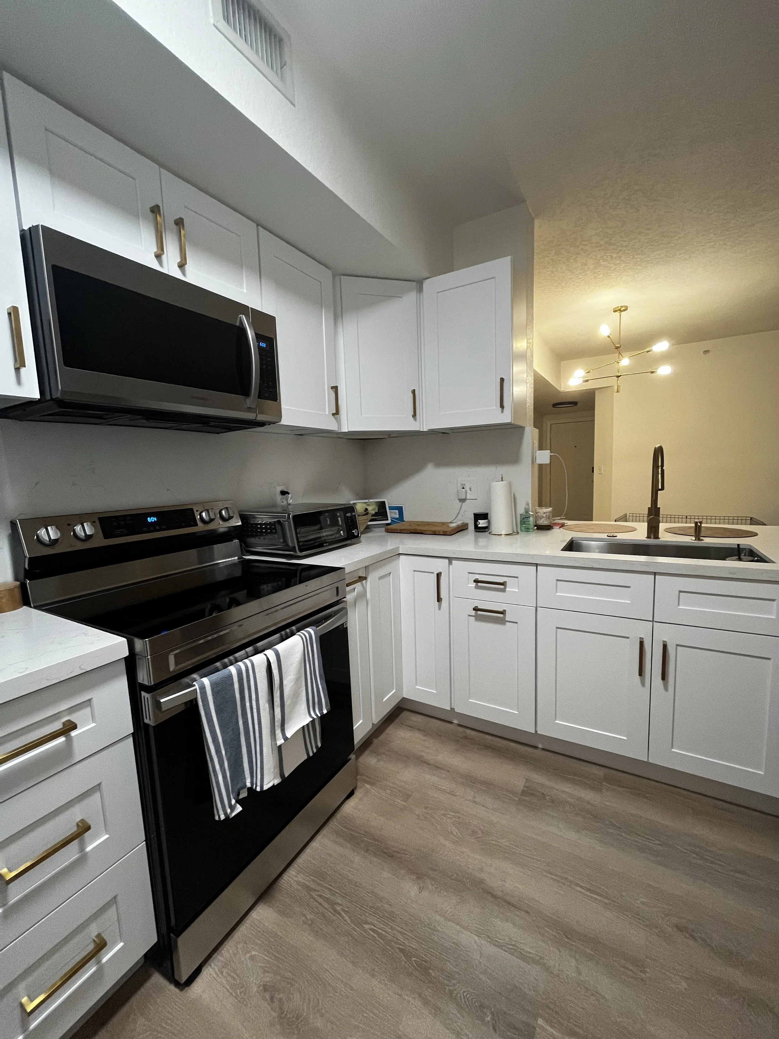 Cocina moderna con gabinetes blancos, estufa de acero inoxidable, microondas sobre la estufa, encimera blanca, lavabo de acero inoxidable, piso de madera clara, y luces decorativas en el techo.