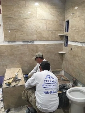 Dos trabajadores en una obra de construcción, uno de ellos con una camiseta que dice 'Zelamari Construction' y un número de contacto, trabajando en la instalación de azulejos en una pared, con herramientas y materiales de construcción alrededor.
