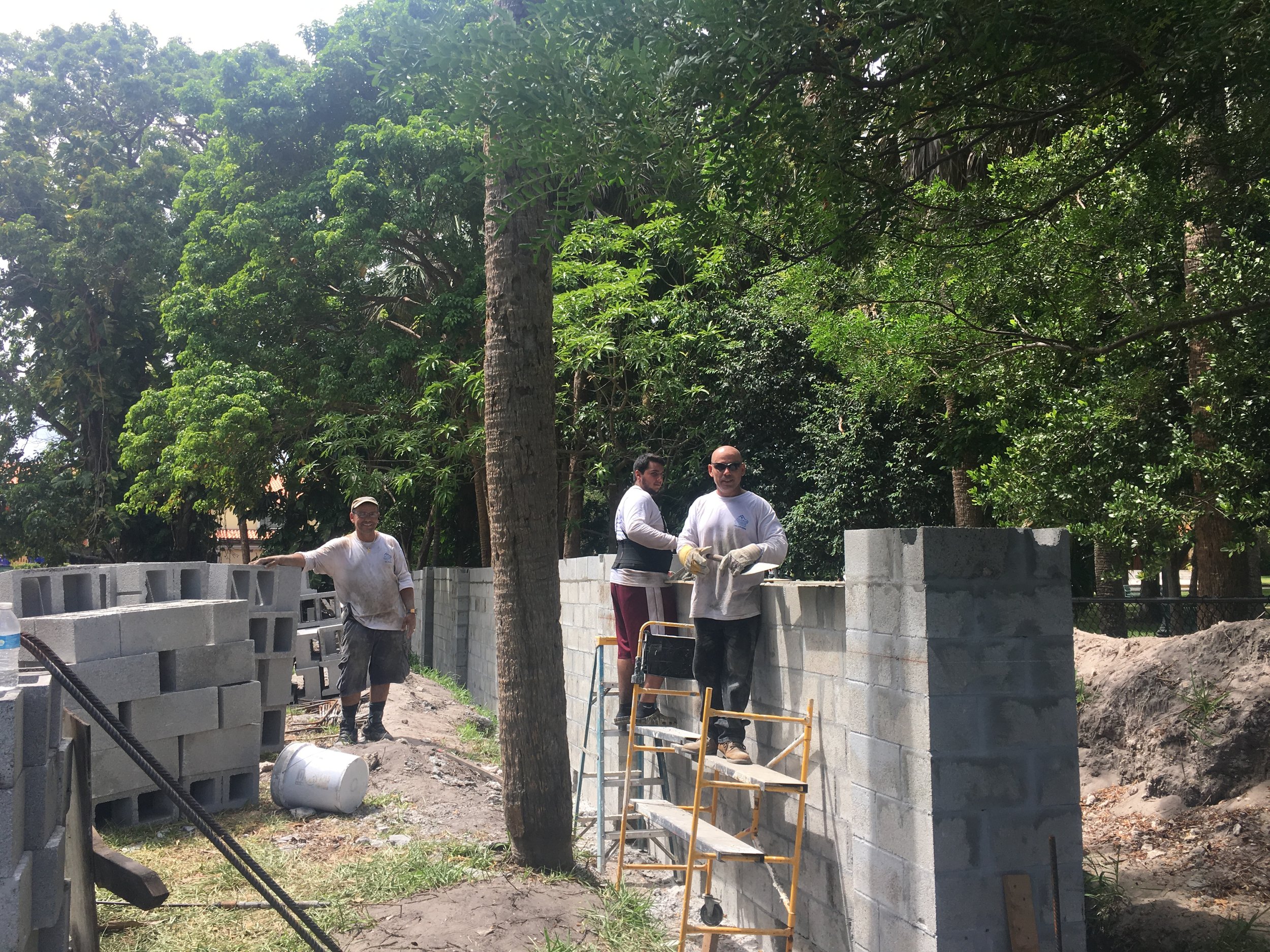 Tres trabajadores construyen una pared de bloques en un área al aire libre con árboles verdes en el fondo.