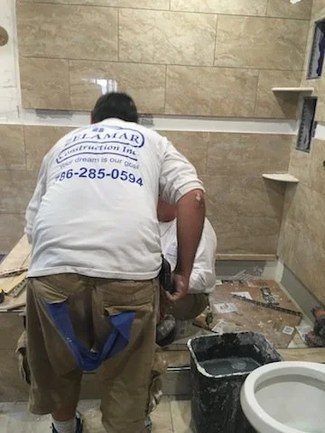 Dos trabajadores instalando azulejos en una pared del baño. Uno de ellos lleva una camiseta blanca con logotipo y teléfono en la espalda, y están rodeados de herramientas y materiales de construcción.