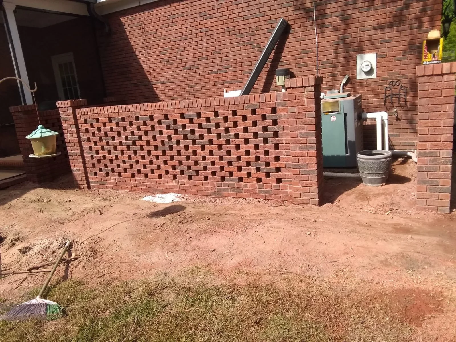 Área de patio con tierra, sin césped, con una pequeña cerca de ladrillos rojos y paredes de ladrillo, con algunos objetos de jardinería, como una escoba y varios recipientes, incluyendo un sistema de ventilación y un tanque de agua.