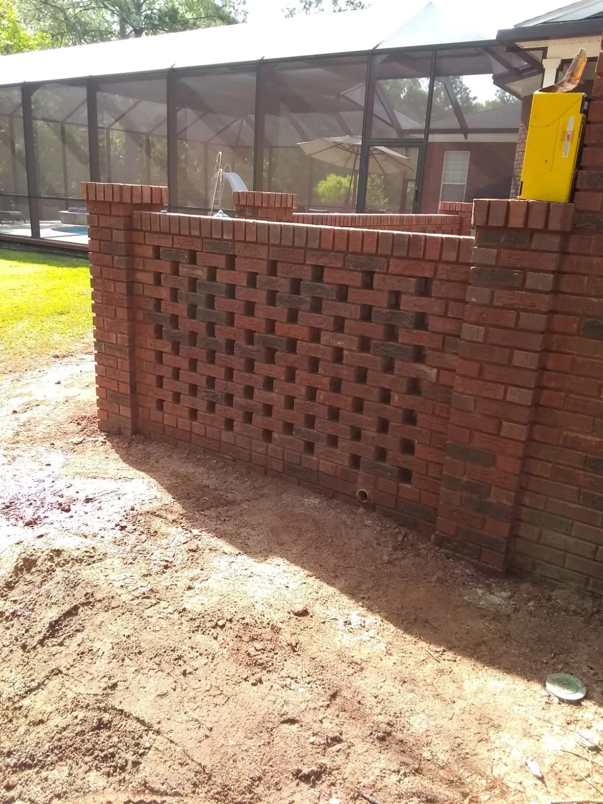 Pared de ladrillos con diseño en el centro, ubicada al lado de una estructura de pantalla de malla para piscina.