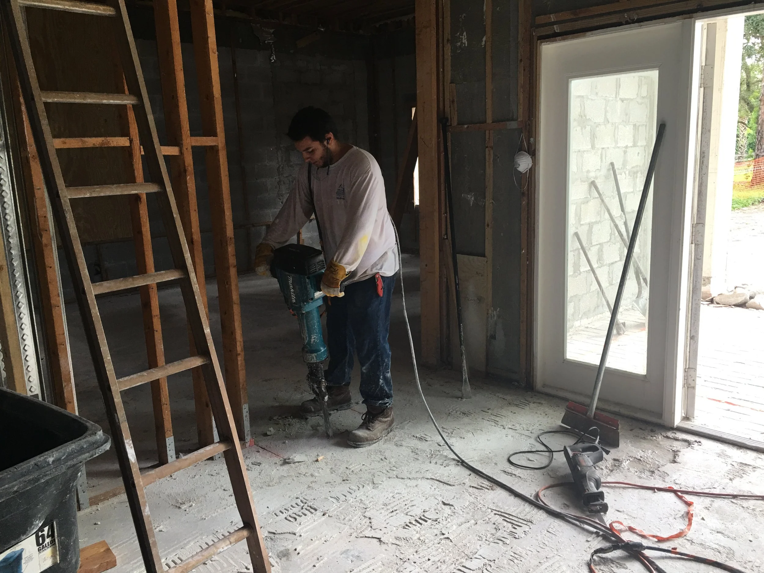 Un hombre trabajando en una construcción interior, usando una herramienta eléctrica de perforación en el suelo de concreto.