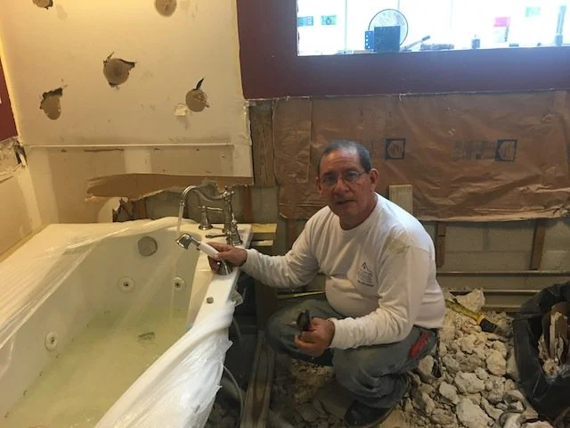Hombre con gafas y camisa blanca arreglando una bañera en una remodelación de baño con paredes con agujeros y escombros.