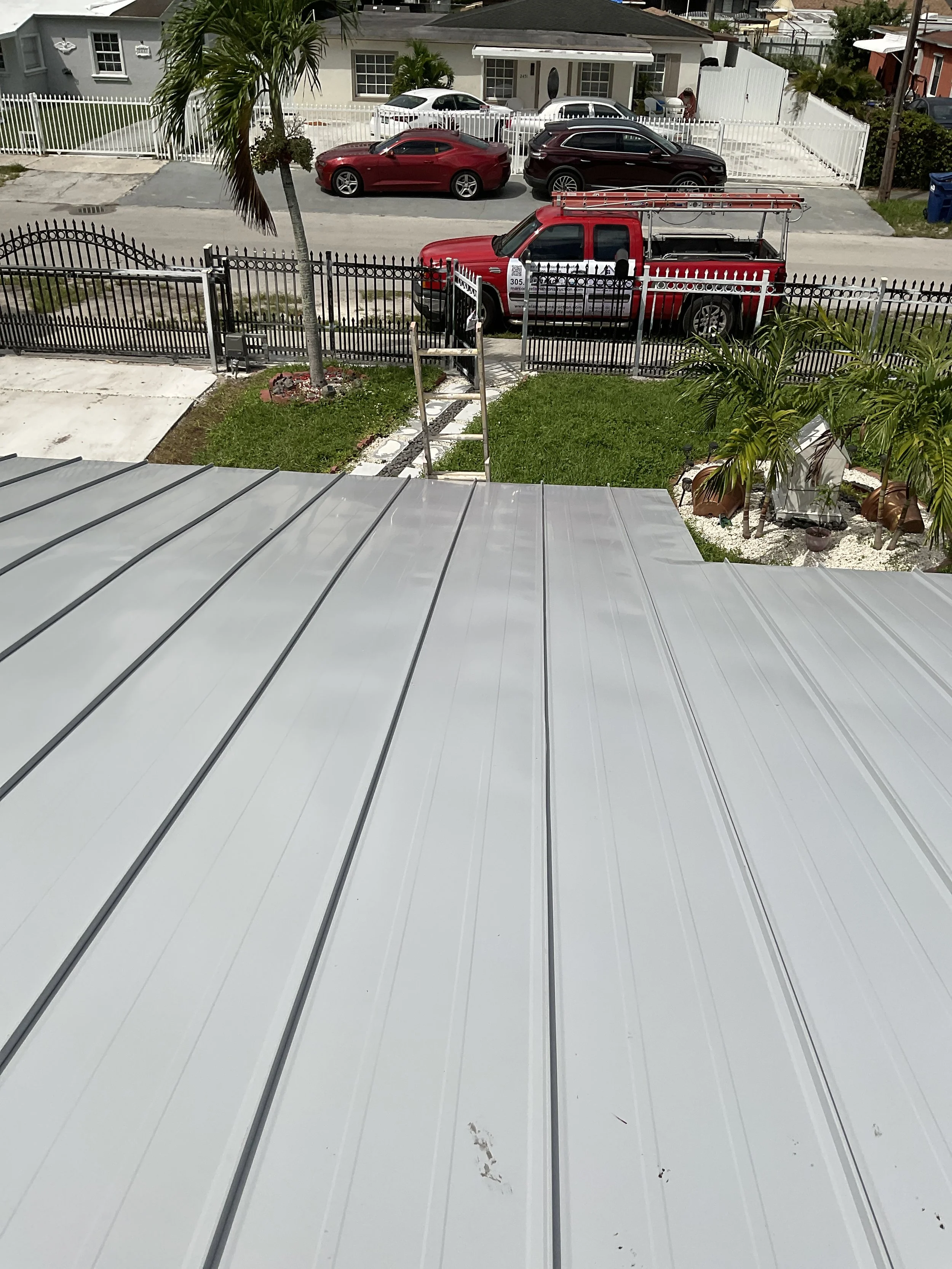 Vista desde una ventana con techo metálico hacia un jardín con arbustos y palmeras, y una calle con varios autos, incluyendo un camión de bomberos estacionado frente a una reja de metal.