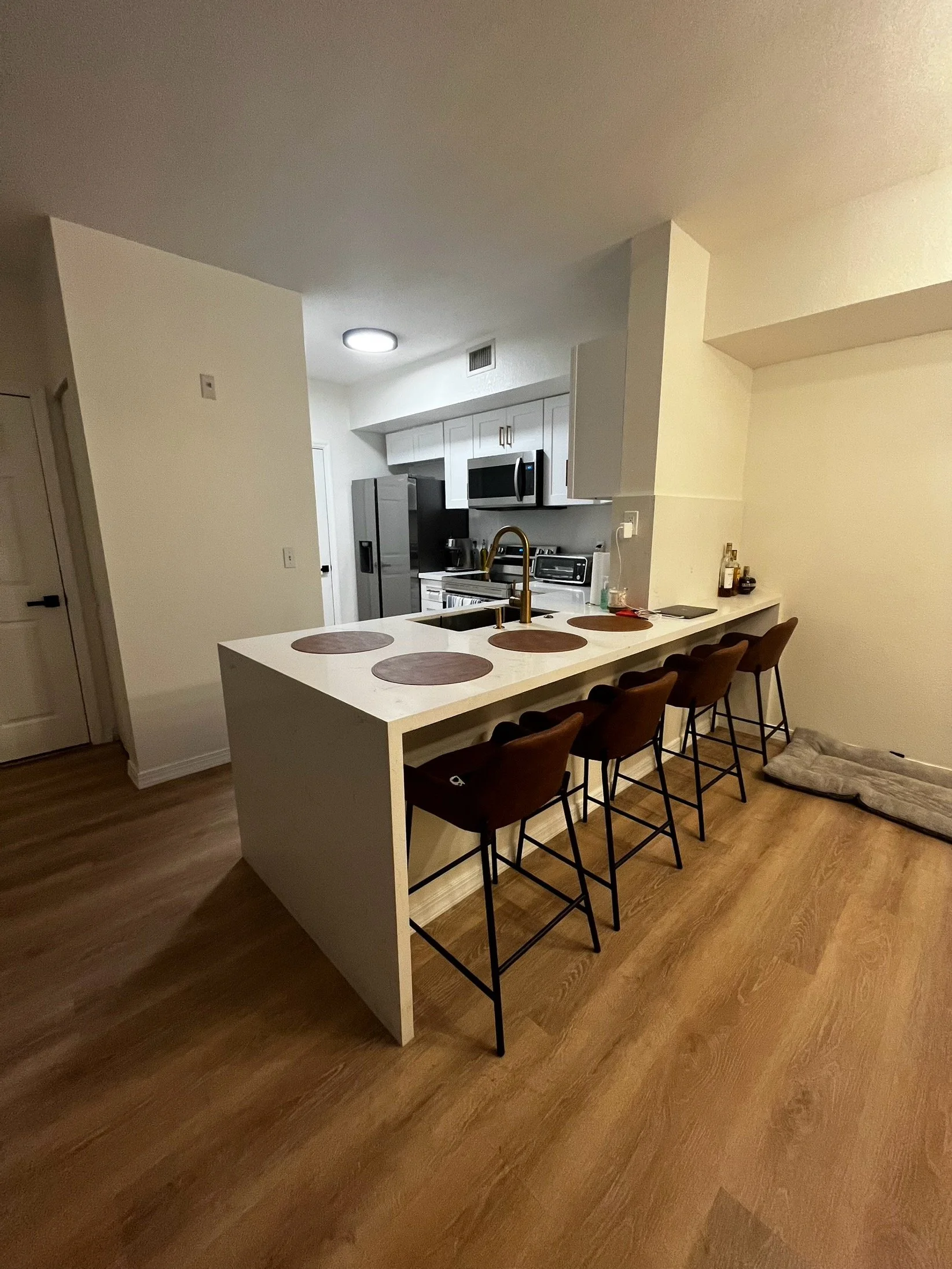 Vista de una cocina moderna con una barra de desayuno blanca, cuatro sillas de asiento marrón y un área con utensilios y botellas en la encimera.
