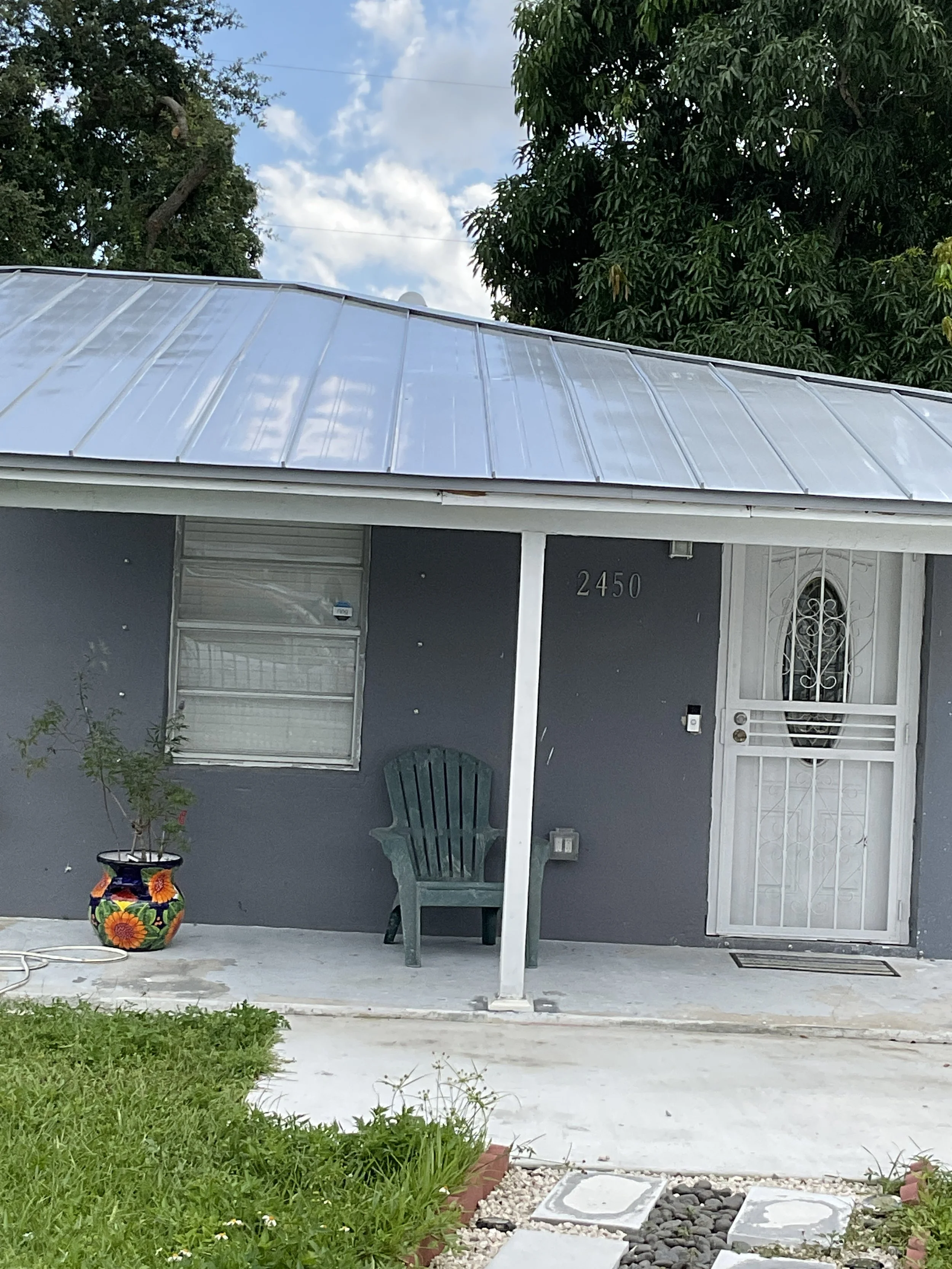 Vista frontal de una casa con paredes grises, puerta metálica blanca y una ventana con reja, en el porche hay una silla de plástico verde y una maceta con planta y flores coloridas.