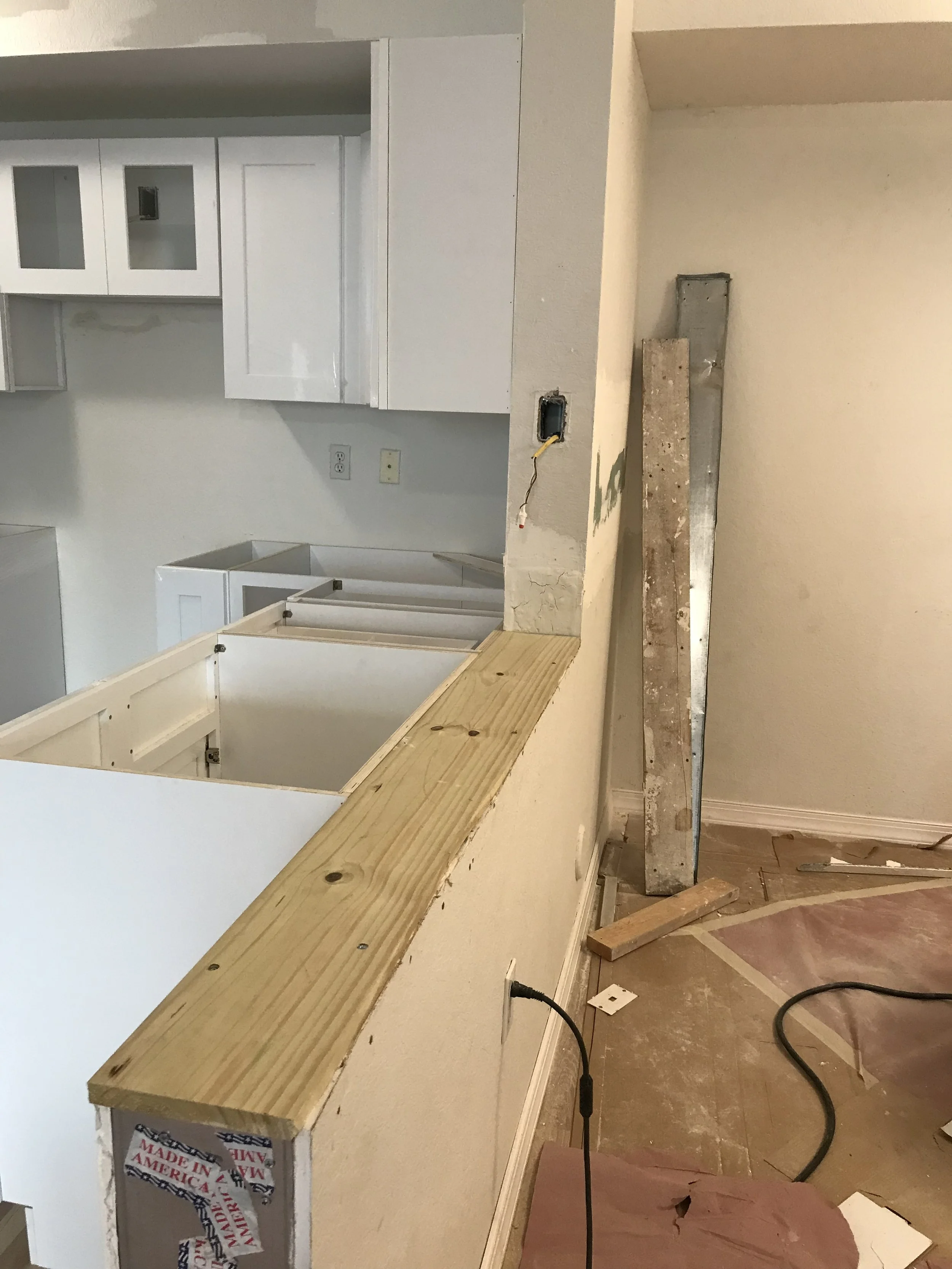 Vista de una cocina en construcción con un muro dividido; en la parte superior hay gabinetes blancos y en la parte inferior hay una encimera de madera sin terminar, con herramientas y materiales de construcción a su alrededor.