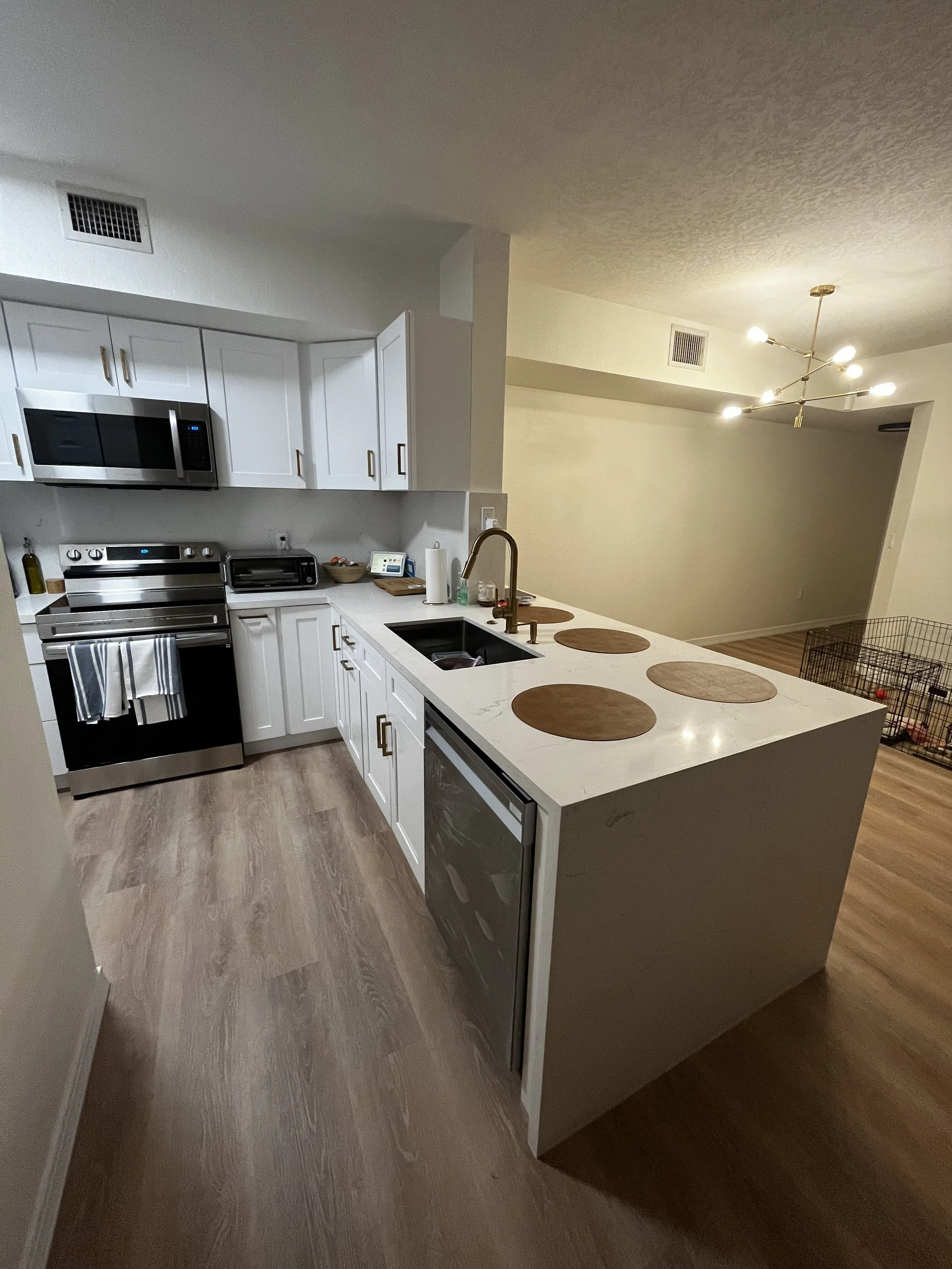 Cocina moderna con electrodomésticos de acero inoxidable, gabinetes blancos y encimera tipo isla con tapetes redondos de madera, en un espacio abierto con iluminación moderna y suelo de madera.