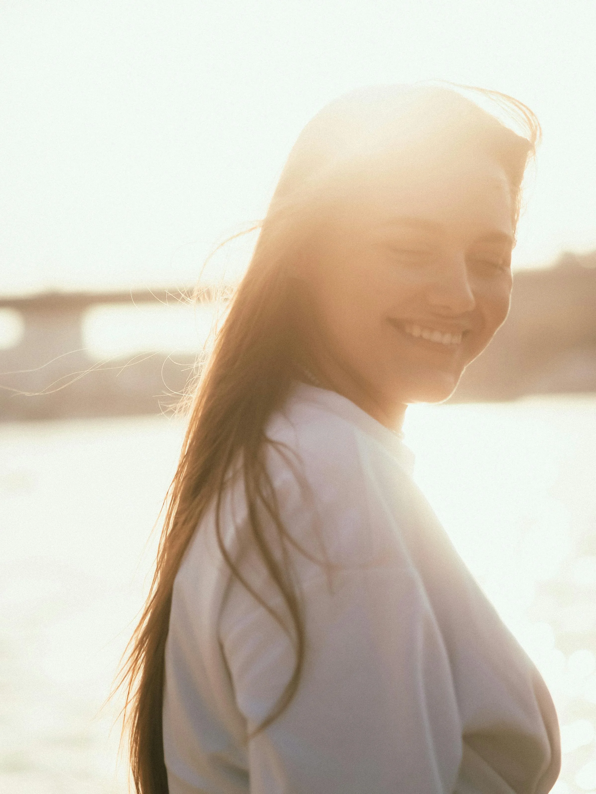 A woman smiling outdoors at sunset, with sunlight backlighting her face and long hair, near a body of water.