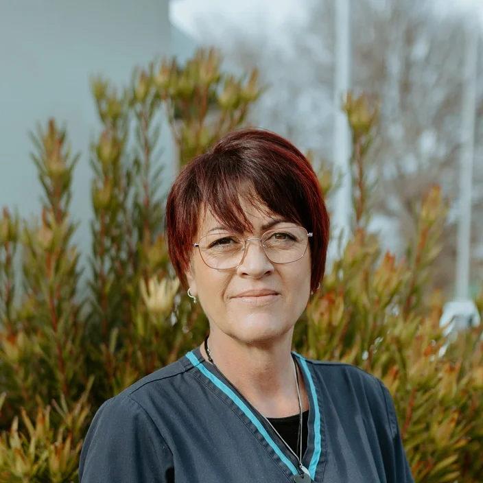 A woman with short red hair, glasses, and earrings stands outdoors with green bushes behind her.