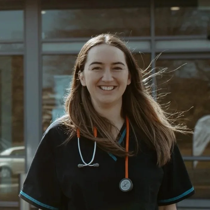 A young woman in medical scrubs smiling outdoors with a stethoscope around her neck.